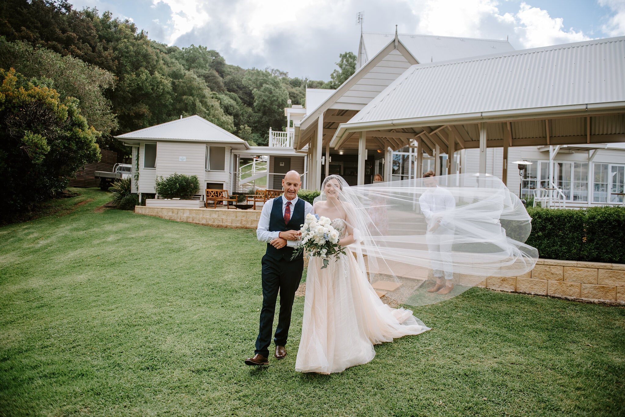 Jamie’s dad walks her across the lawn to the ceremony, her veil streaming in the breeze with Maleny Manor and its pavilion behind.