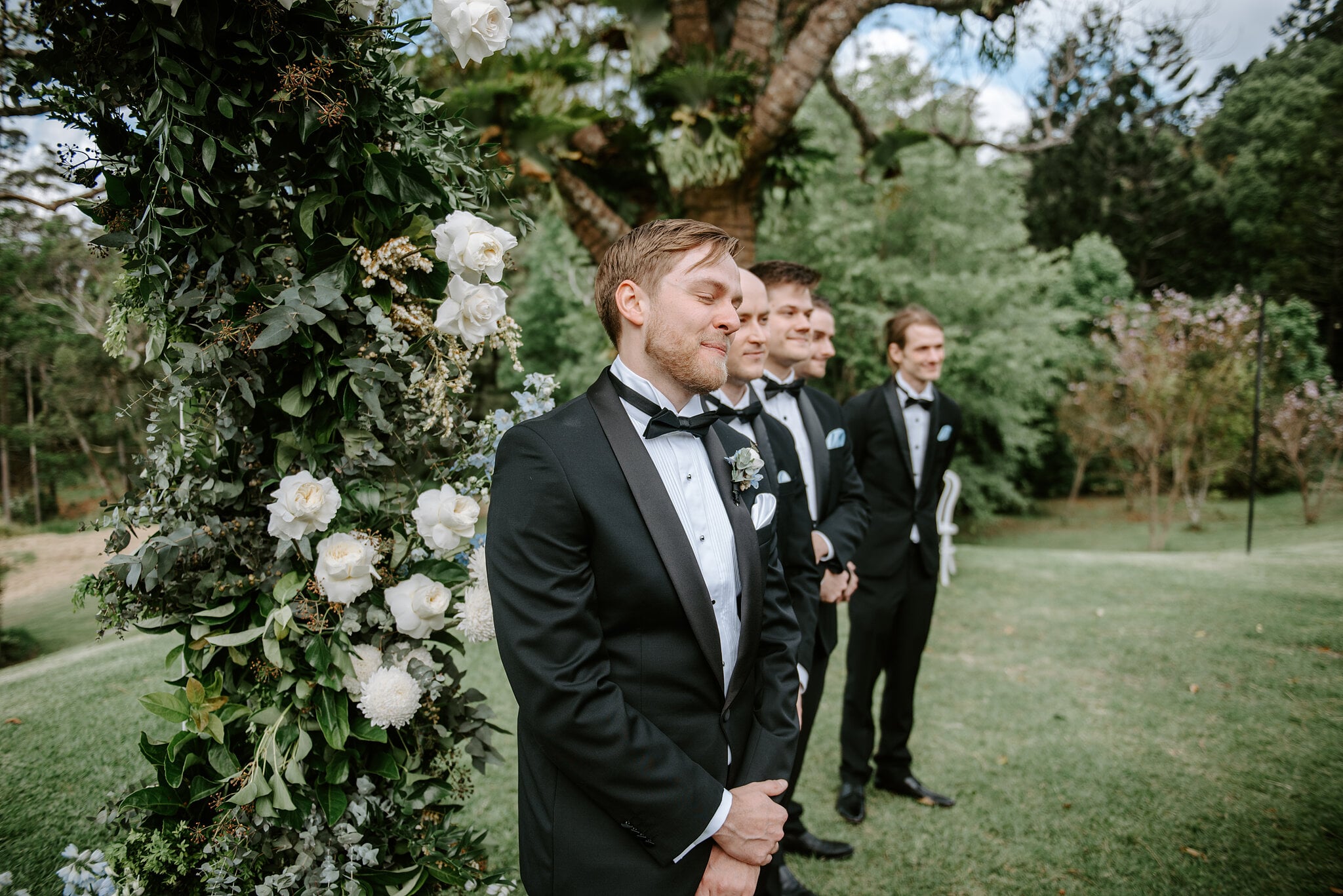 Tim waits beside the white rose arch with his groomsmen, eyes closed, taking a deep breath.
