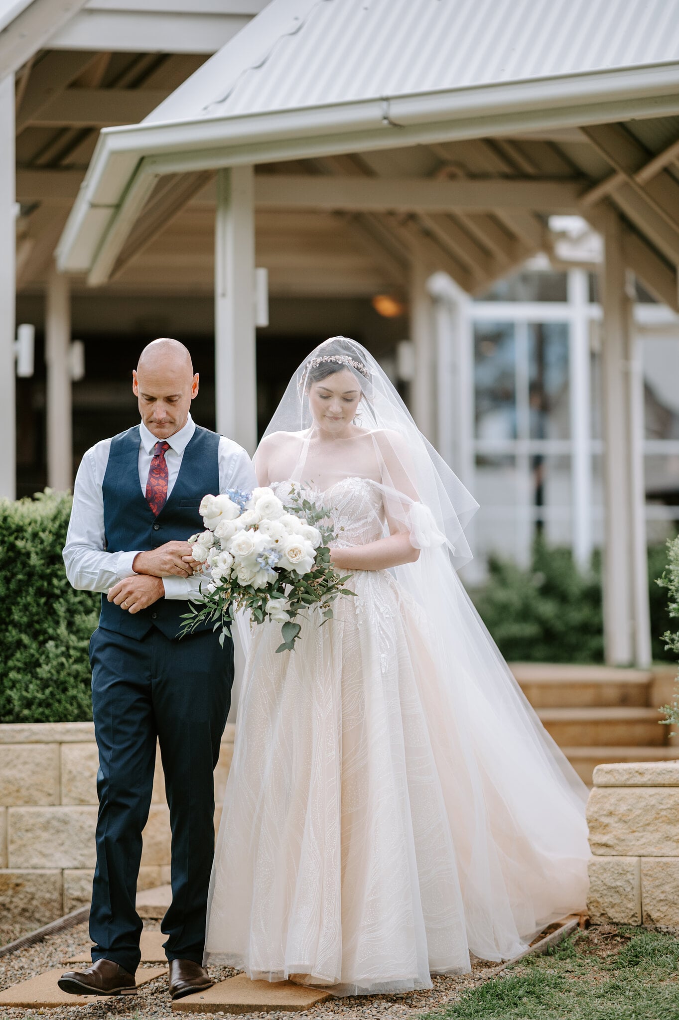 Jamie comes down the sandstone steps beside the pavilion on her dad’s arm, her veil over her face.