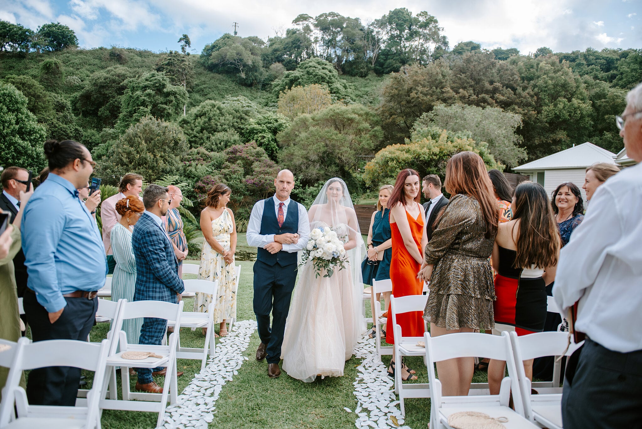 Guests stand beside white chairs as Jamie’s dad walks her down a petal aisle under the green hills of Maleny.