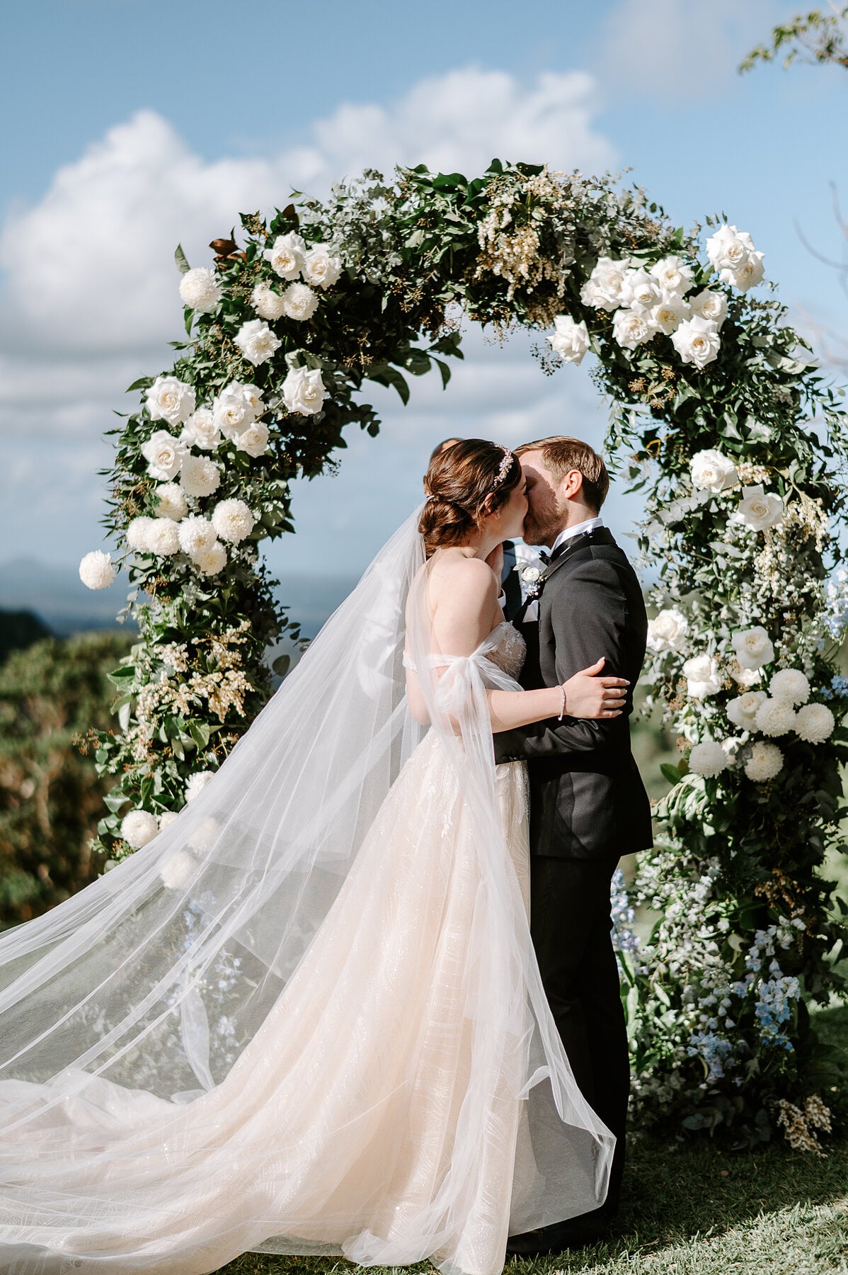 Jamie and Tim share their first kiss under the white rose arch against a blue sky.