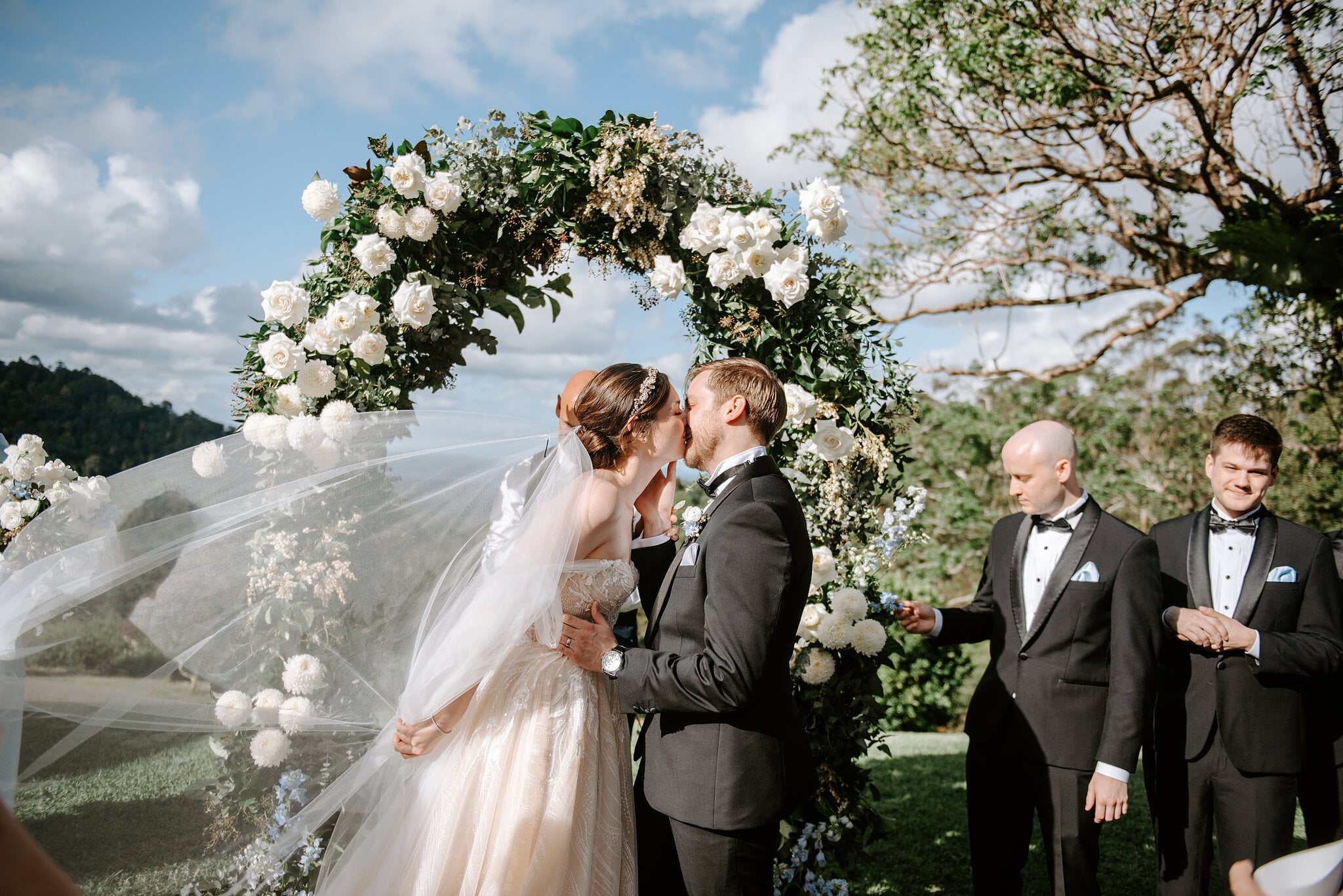 Jamie and Tim kiss again at the arch as her veil sweeps sideways and the groomsmen grin.