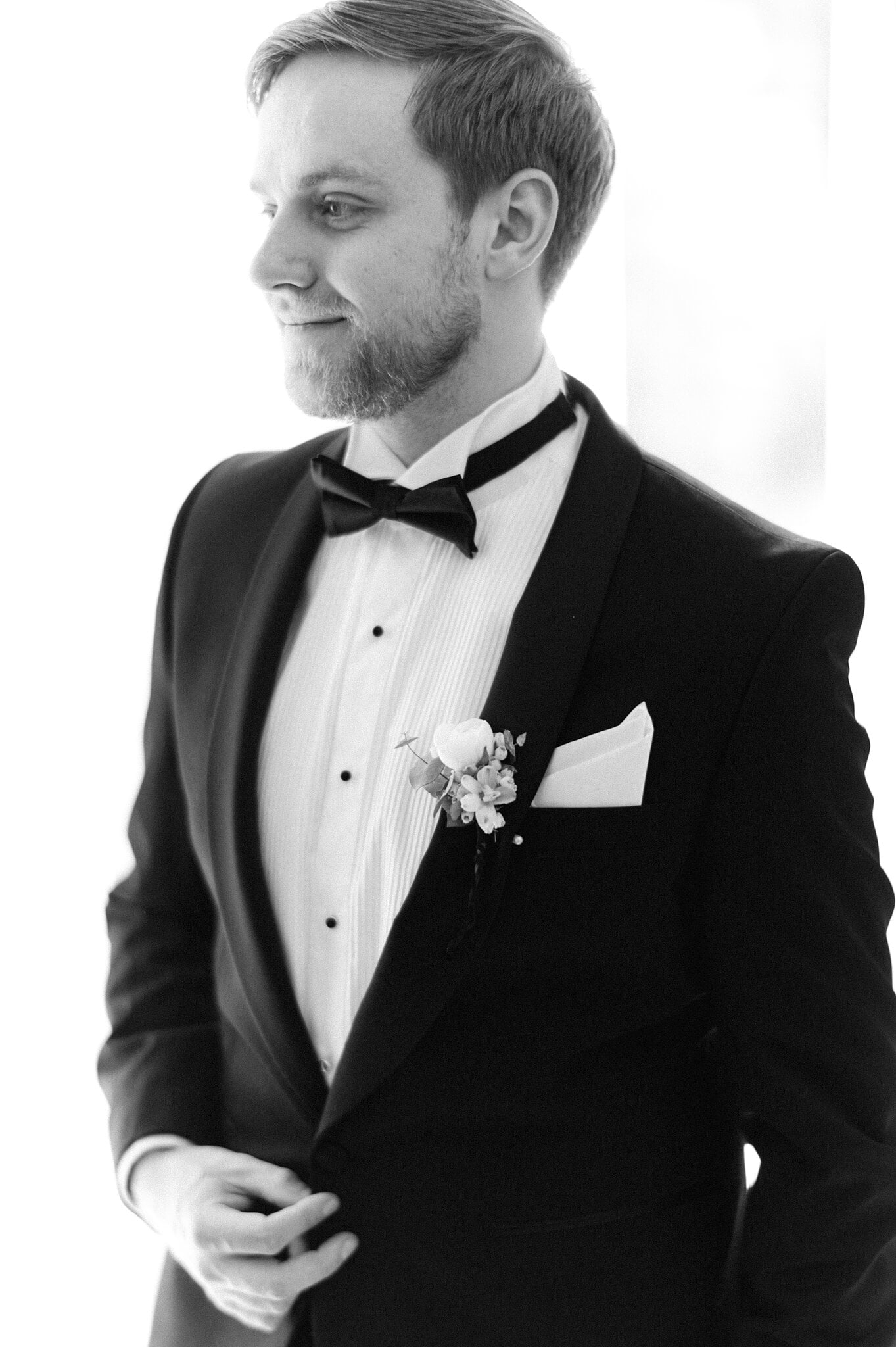 Tim in a black tuxedo and bow tie looks out of a bright window at Maleny Manor, a white rose pinned to his lapel.
