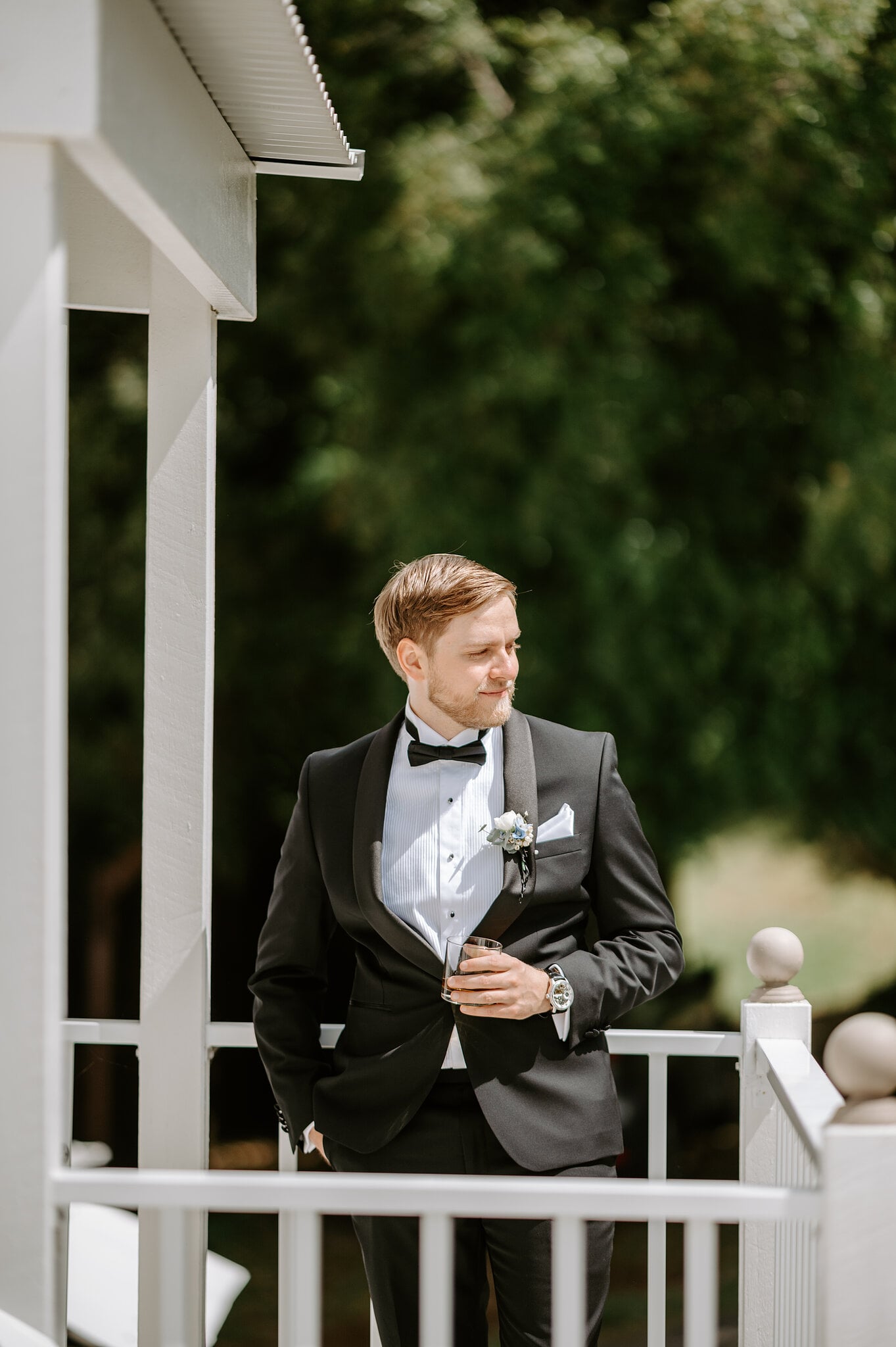 Tim leans on the white veranda railing at Maleny Manor with a glass of whisky, rainforest trees behind him.