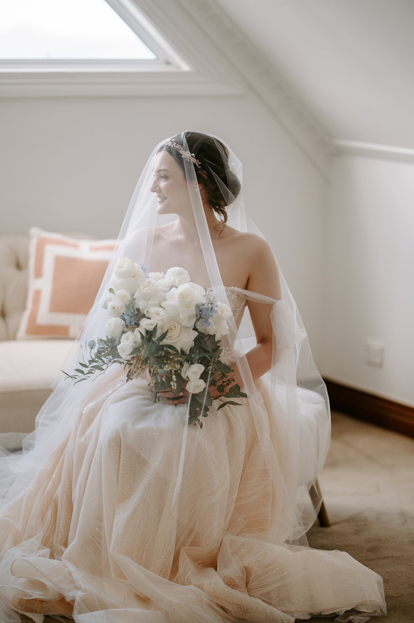 Jamie sits on a cream chaise in her veil, holding white roses and soft blue flowers.