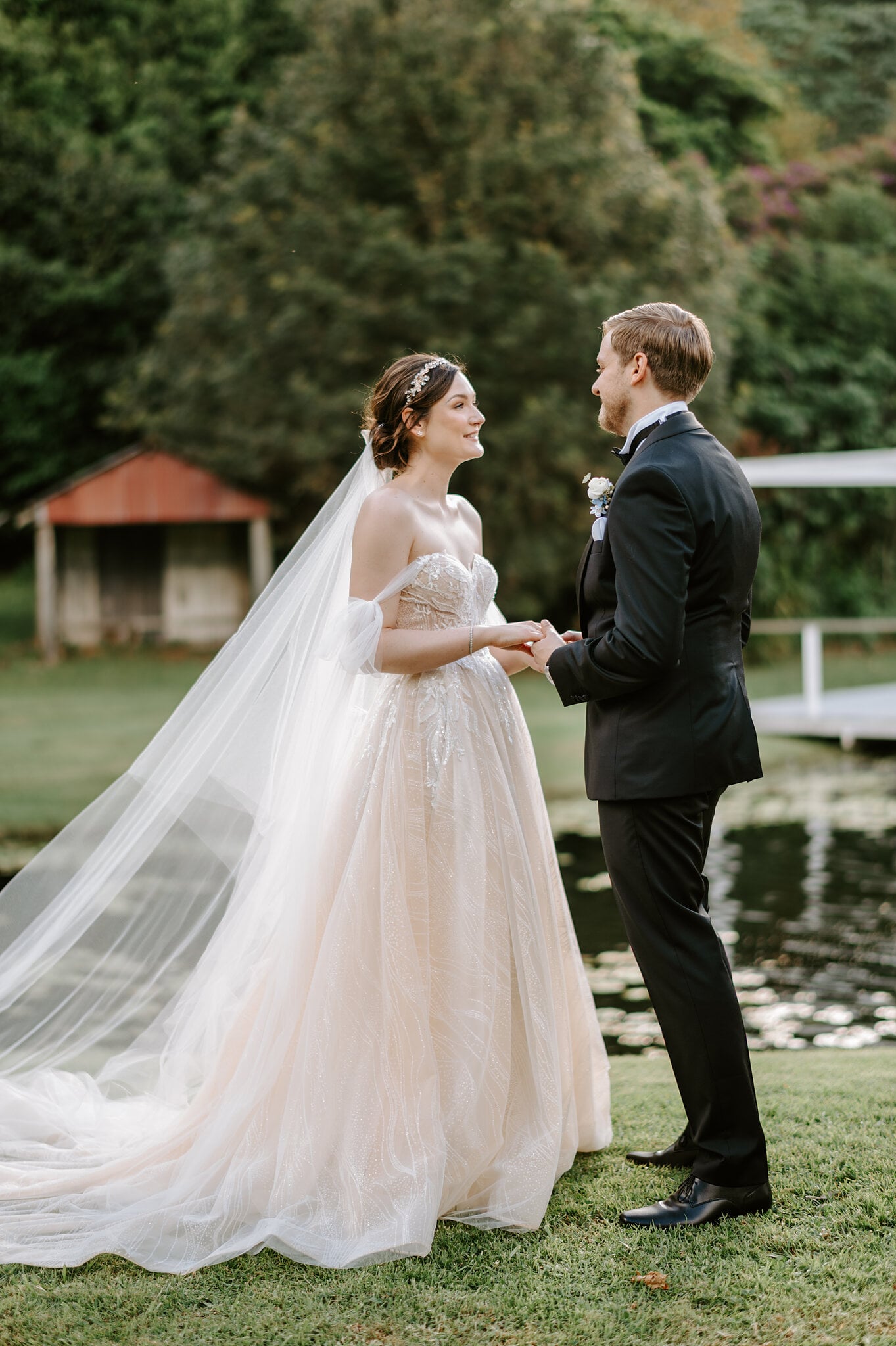 Jamie and Tim hold hands beside the pond at Maleny Manor, a red-roofed boat shed and jetty behind them.