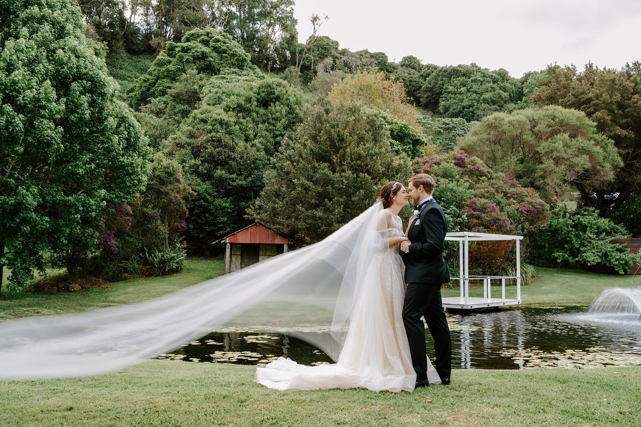 Tim kisses Jamie by the pond as her long veil floats across the lawn, a fountain playing behind them.
