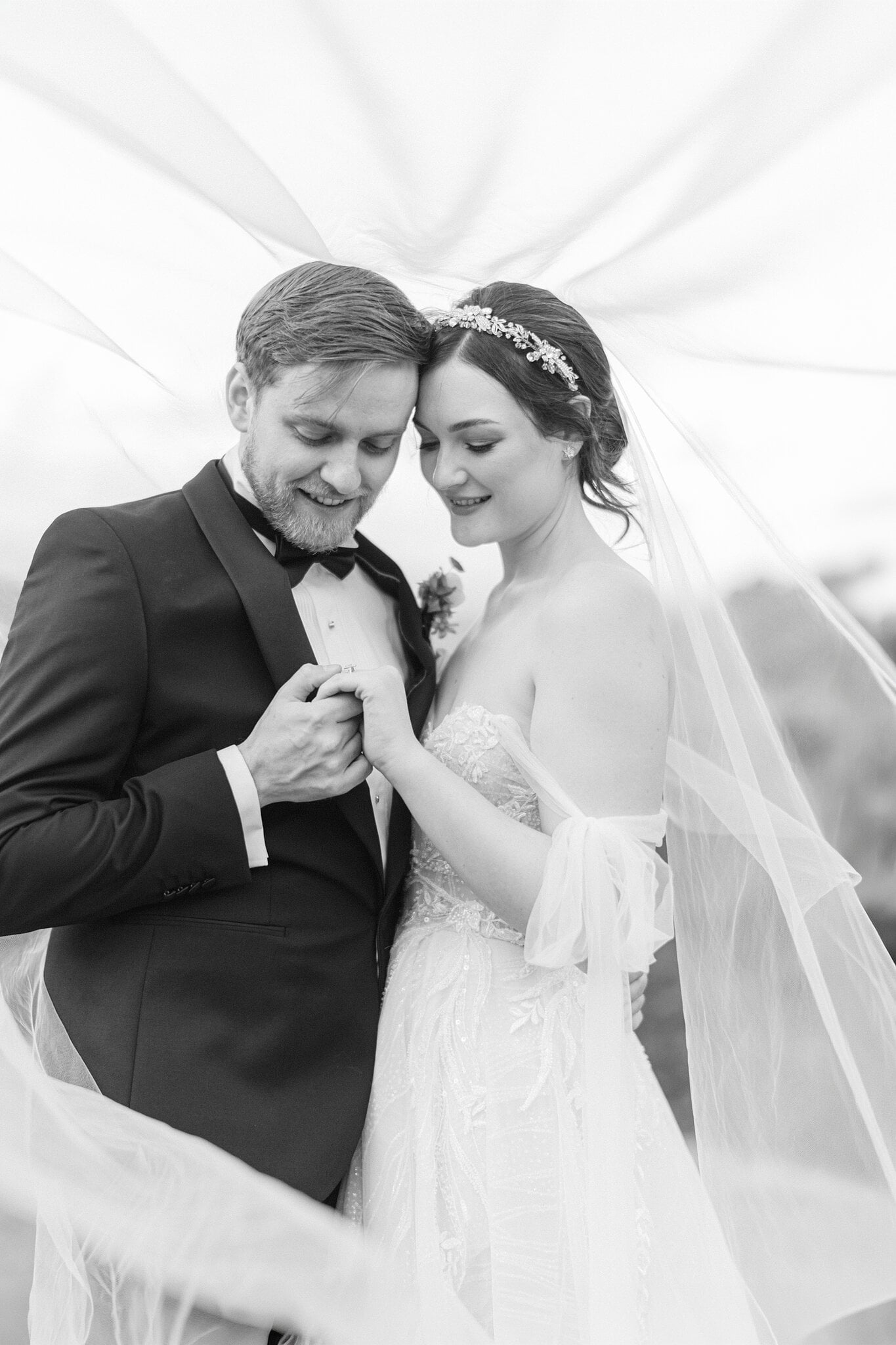 Jamie and Tim hold hands under her veil, foreheads close together, in black and white.
