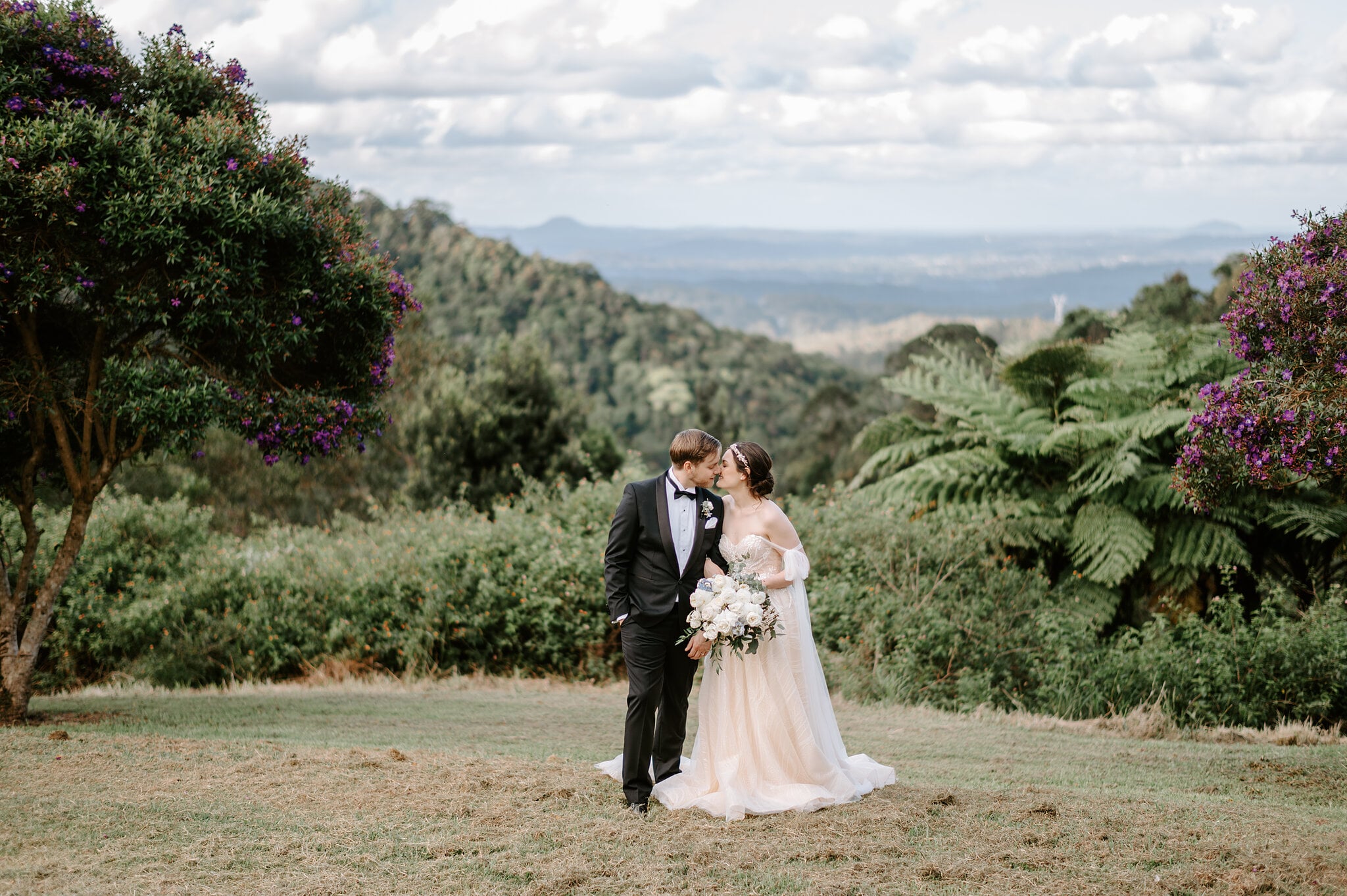 Jamie and Tim kiss on the hilltop at Maleny Manor, the Sunshine Coast hinterland rolling out to the coast behind them.