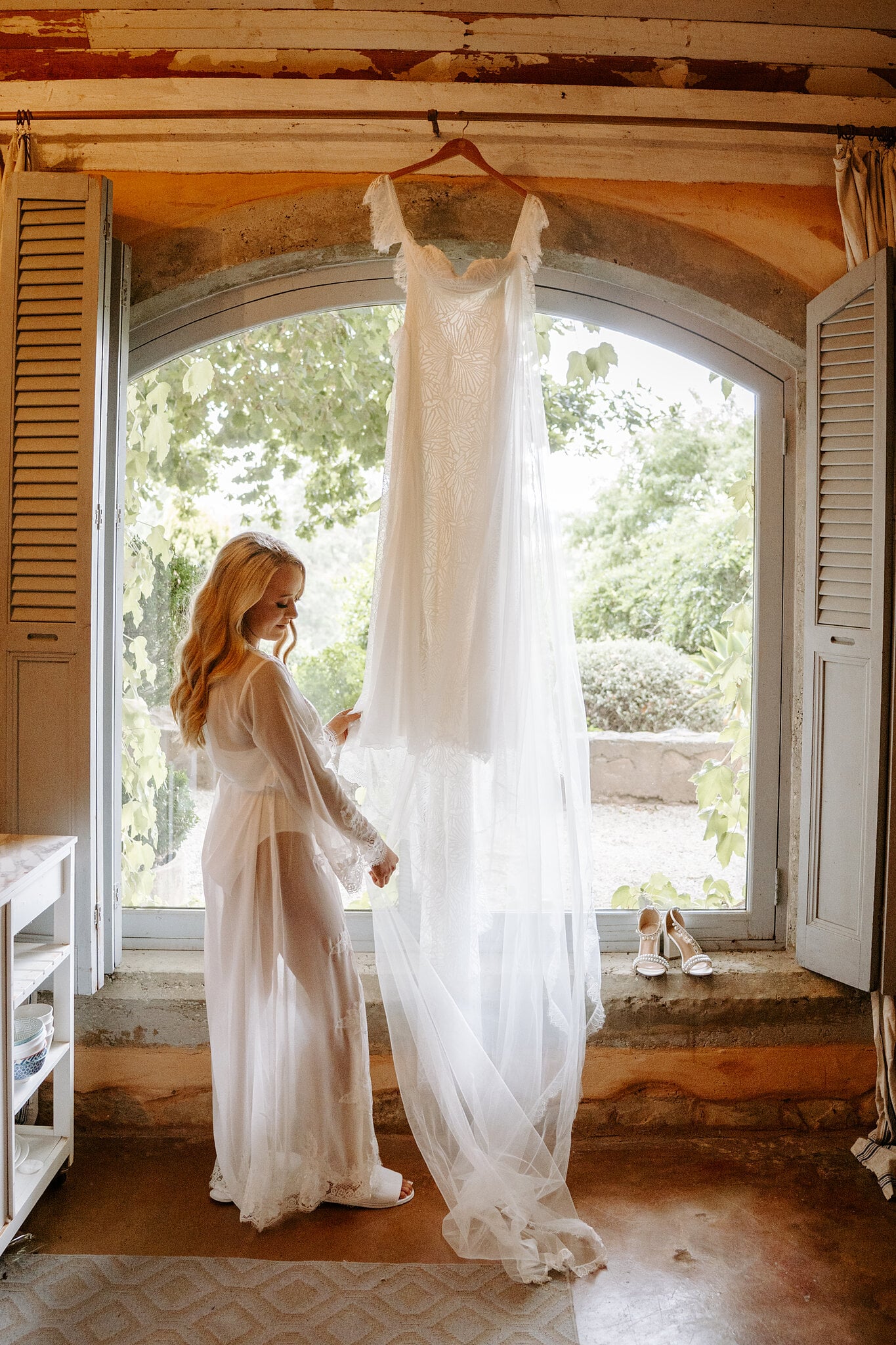 Samantha in a sheer lace robe beside her gown hanging in the arched cottage window.