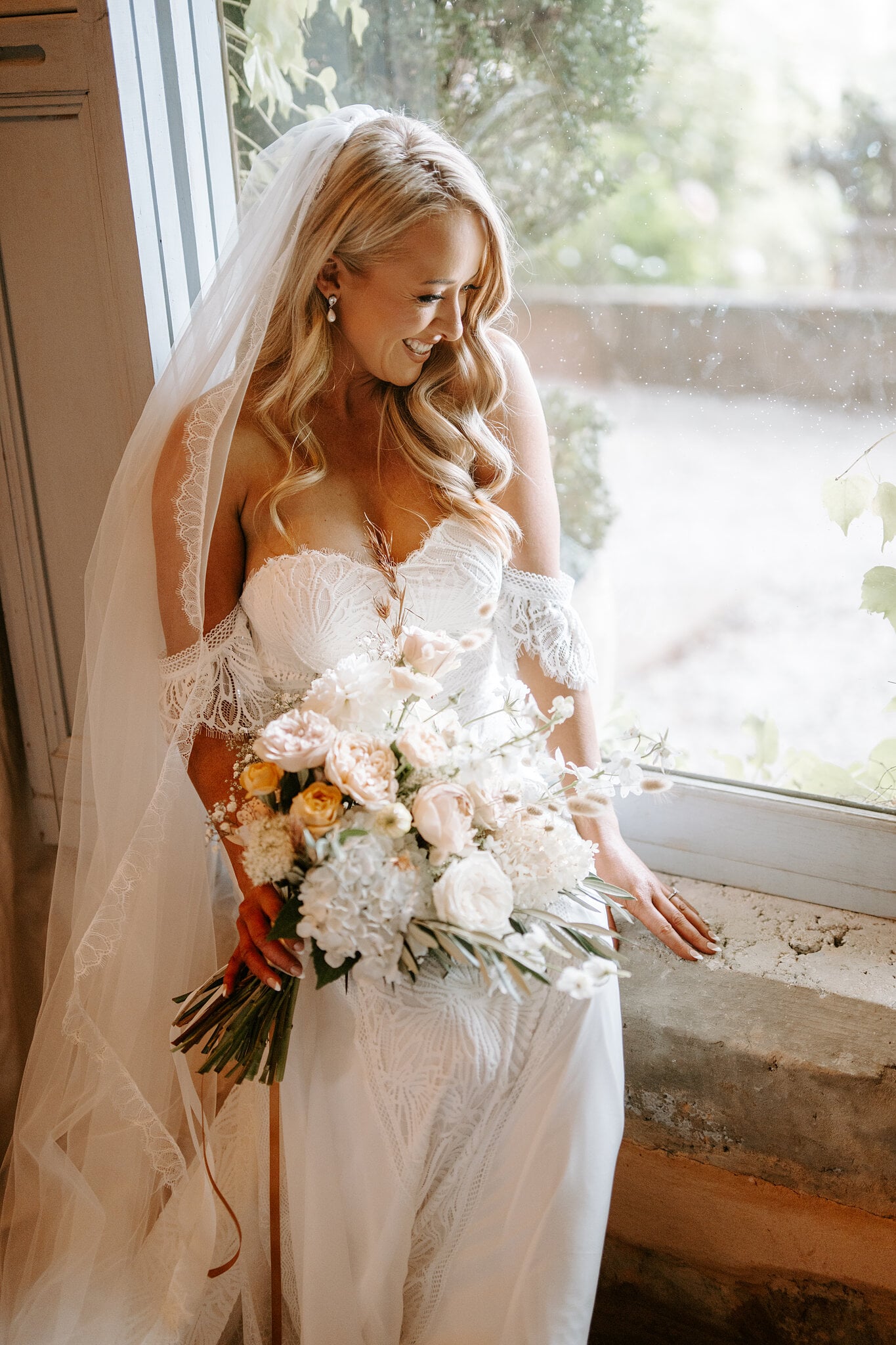 Samantha smiles down at her bouquet by the window, veil falling over her shoulders.