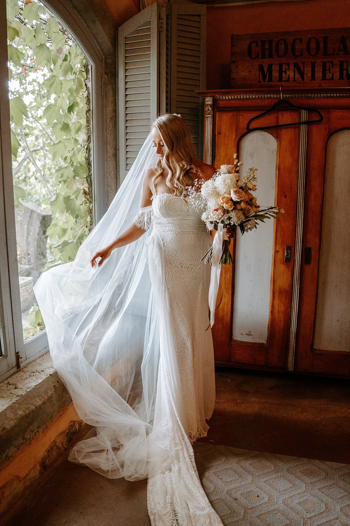 Samantha swishes her veil in the cottage beside an old timber wardrobe and shuttered window.