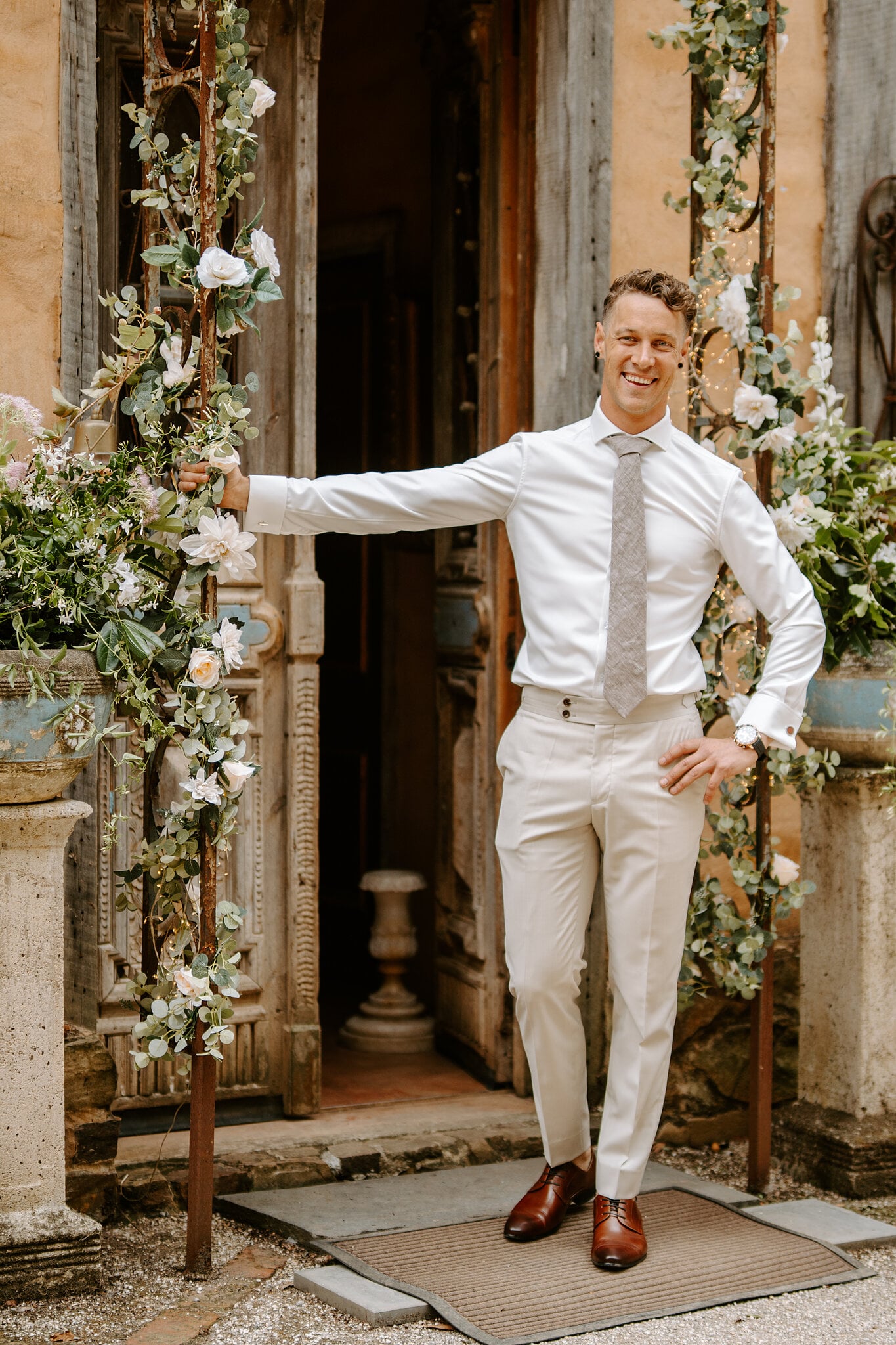 Harrison grins in his shirt and tie at a carved timber doorway framed with roses.