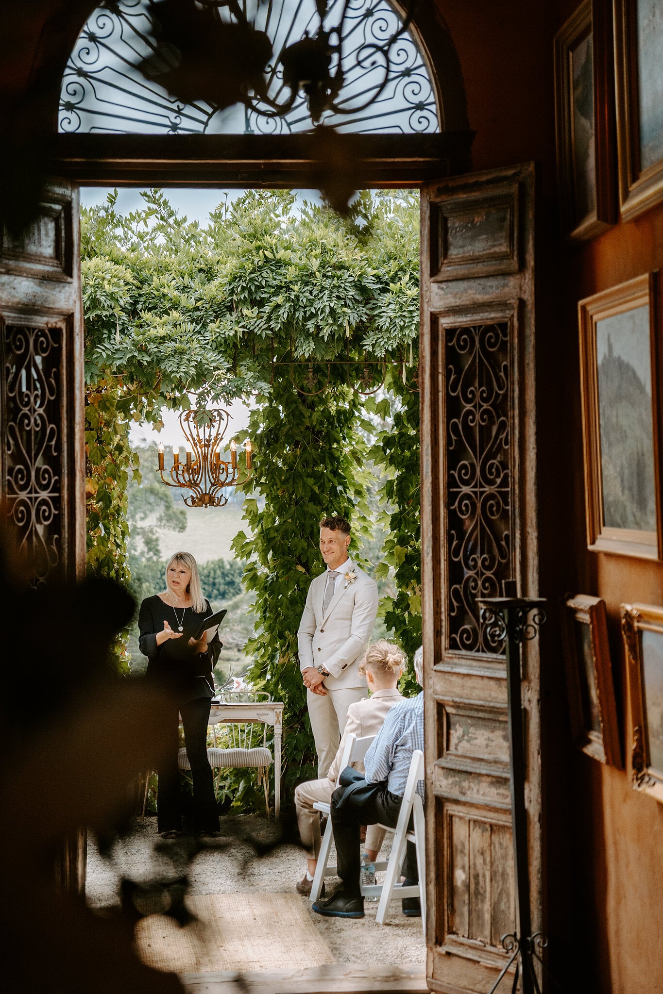 Through carved doors and an iron fanlight, Harrison waits with the celebrant under the vines.