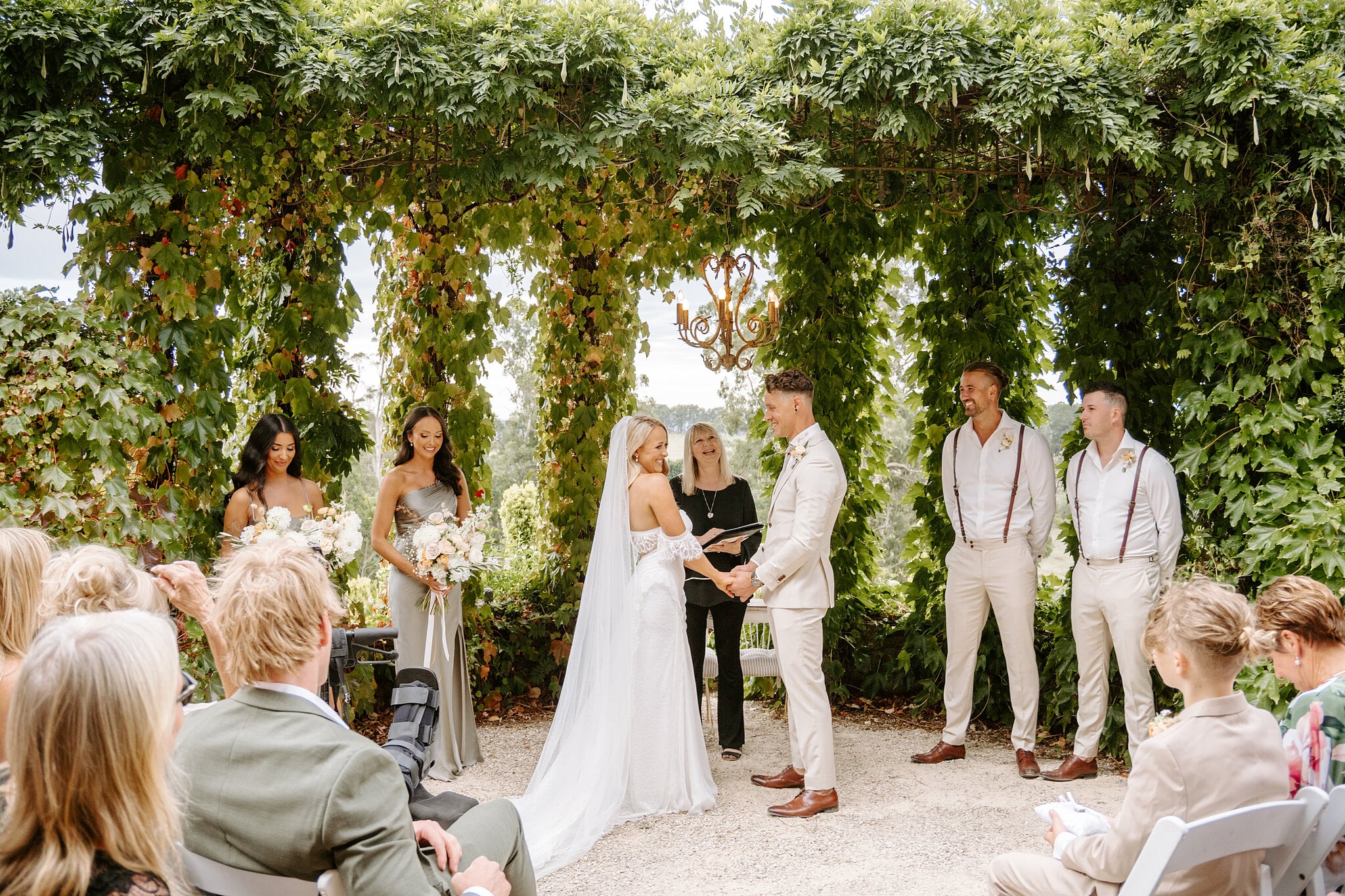 Samantha and Harrison hold hands for their vows under the vine arbour, bridal party either side.