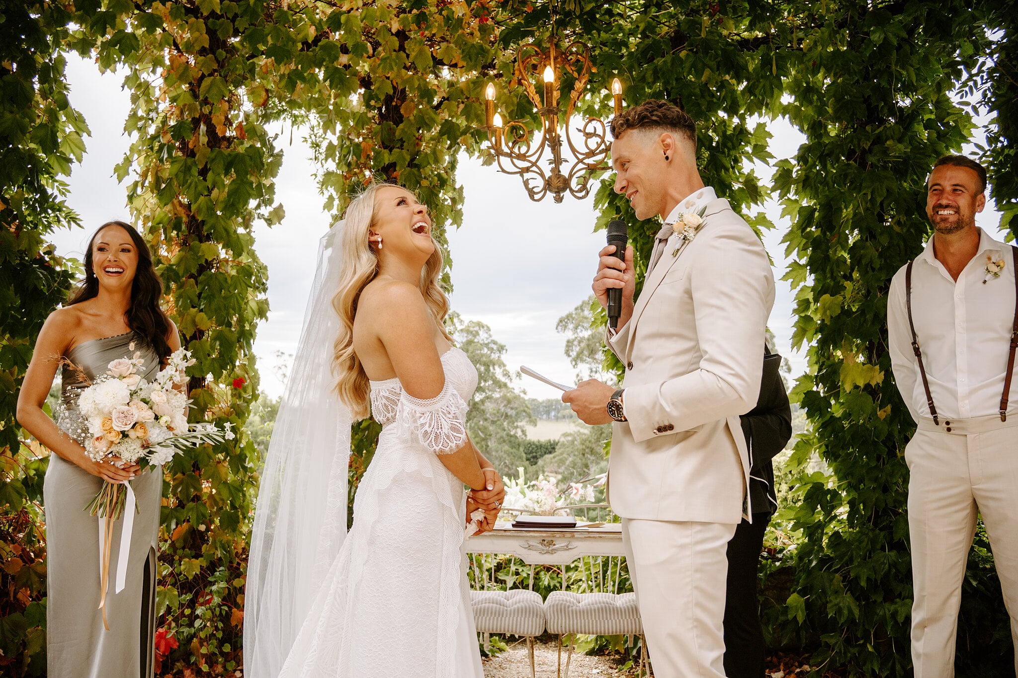 Samantha bursts out laughing as Harrison reads his vows under the arbour chandelier.