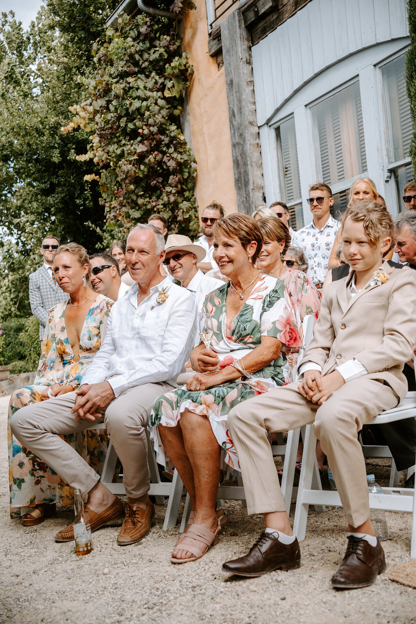 Guests in florals and a page boy in a beige suit laugh along during the vows.