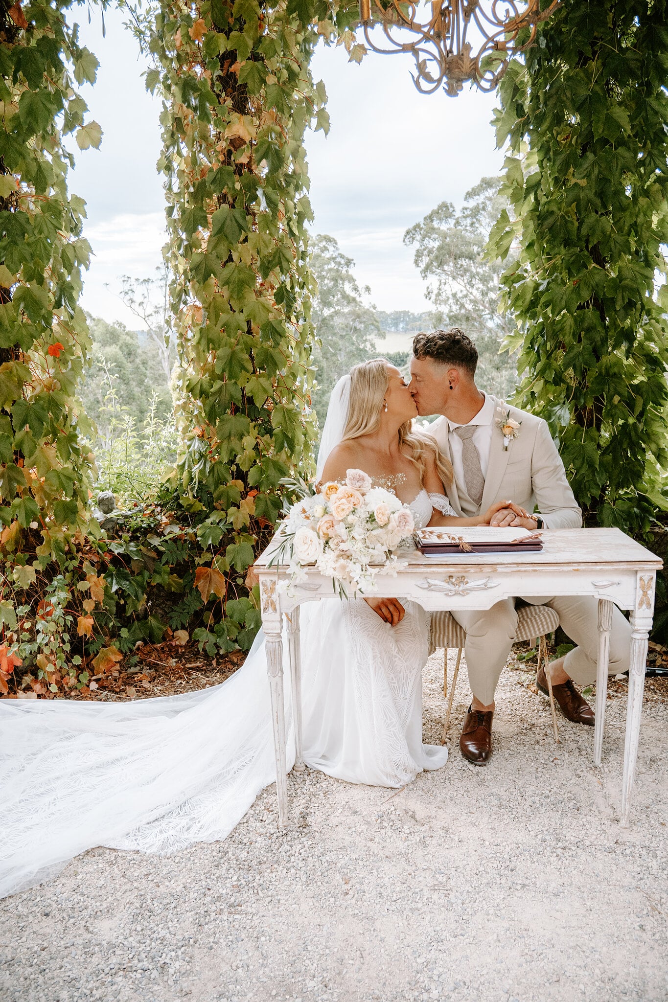 Samantha and Harrison kiss at the signing table under the vines, hills rolling away behind them.