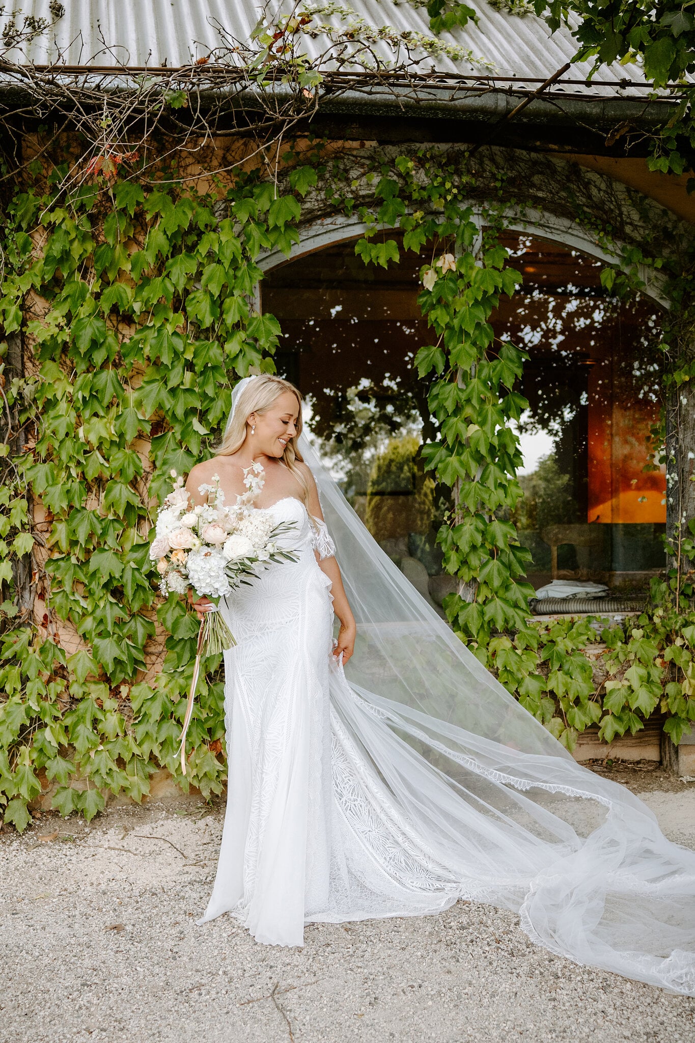 Samantha stands before the ivy-covered arched window of the cottage, her veil pooling on the gravel.