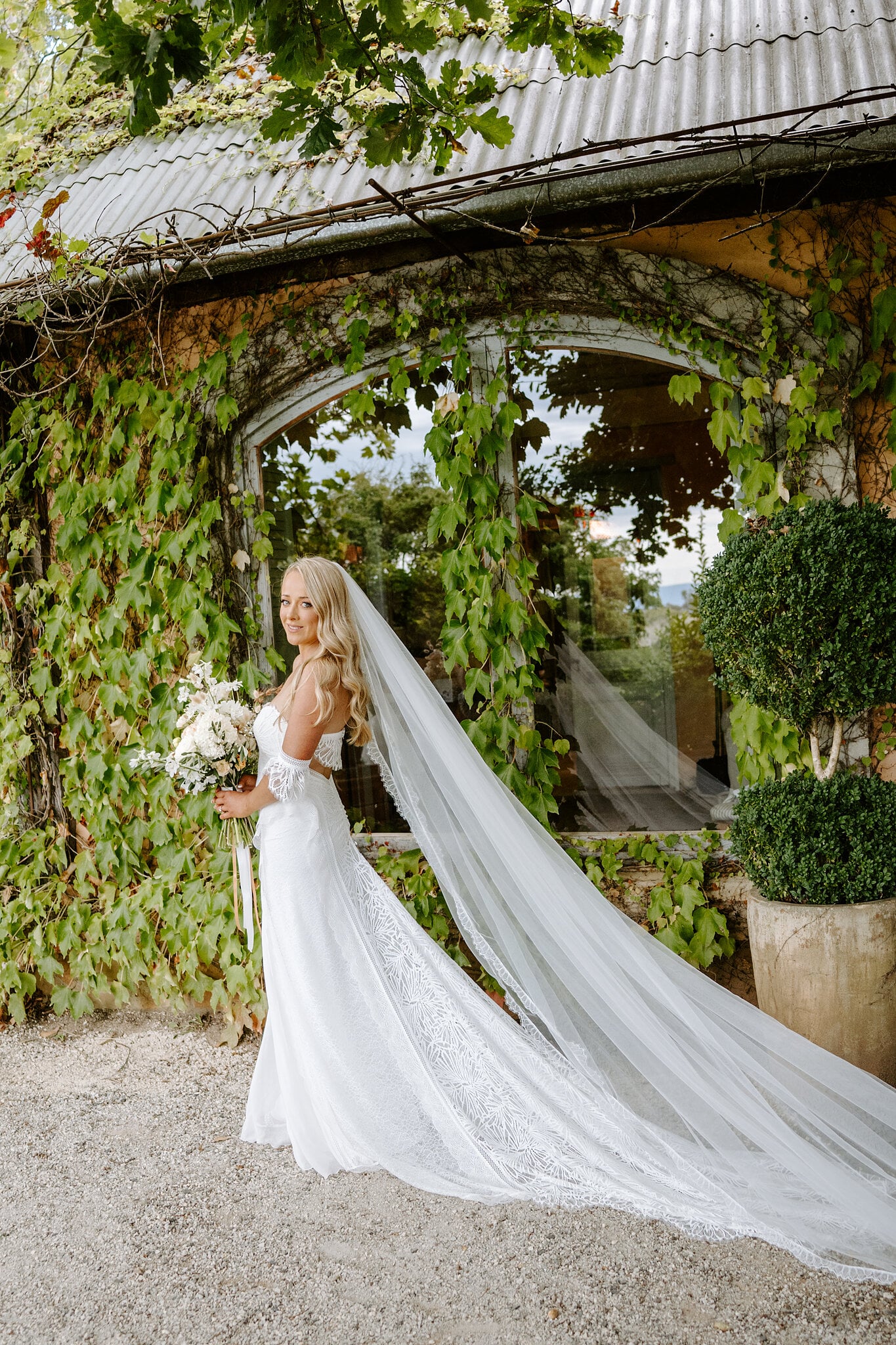 Samantha glances back over her shoulder, her cathedral veil sweeping past the ivy and clipped topiary.