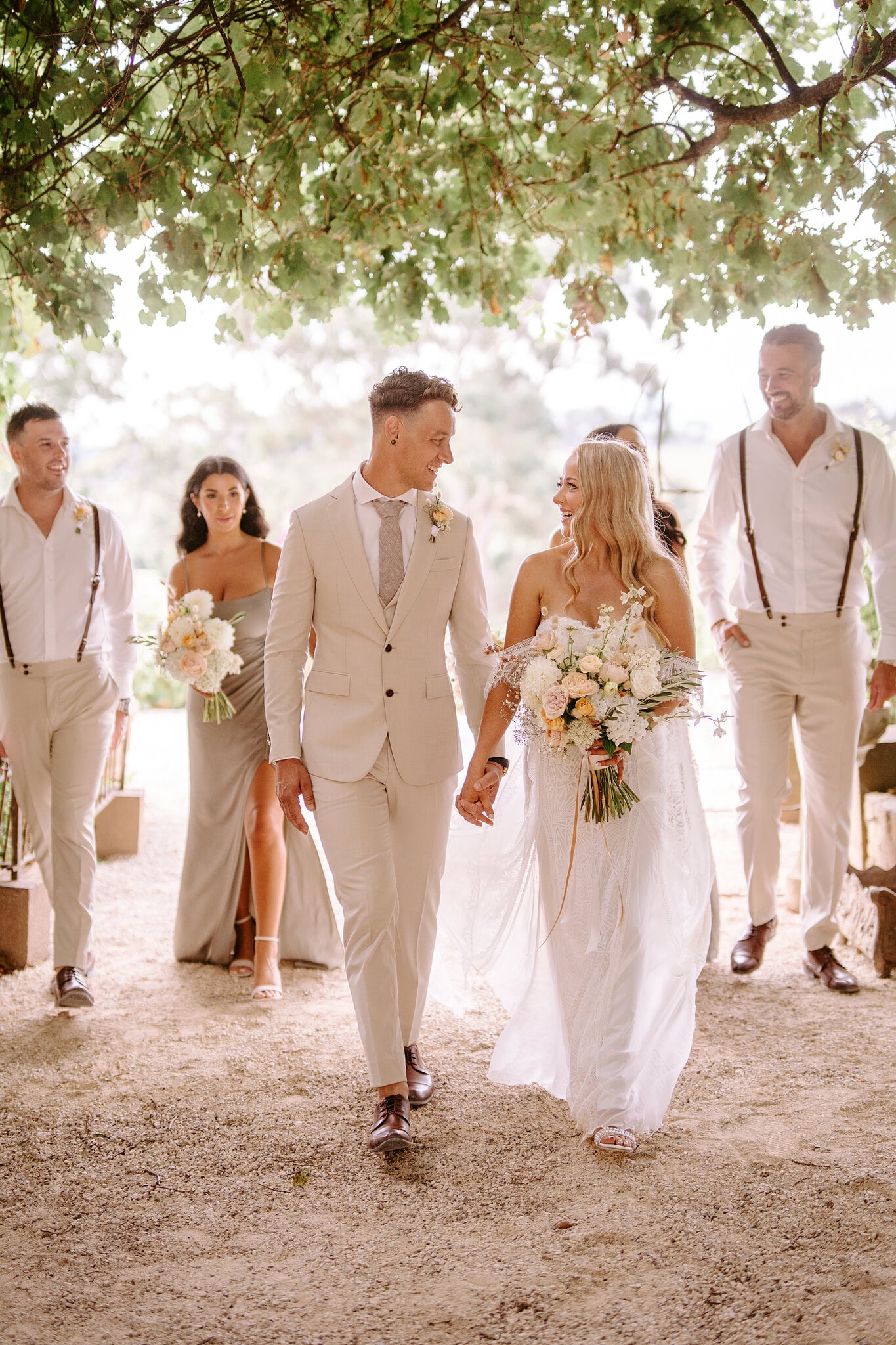 Samantha and Harrison lead their bridal party under a shady tree canopy, hand in hand.