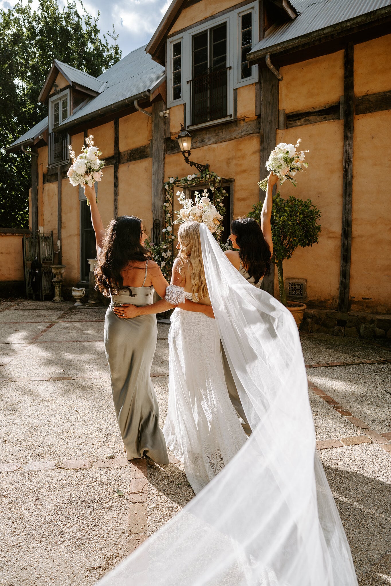 Samantha and her bridesmaids walk off arm in arm, bouquets raised and veil trailing.