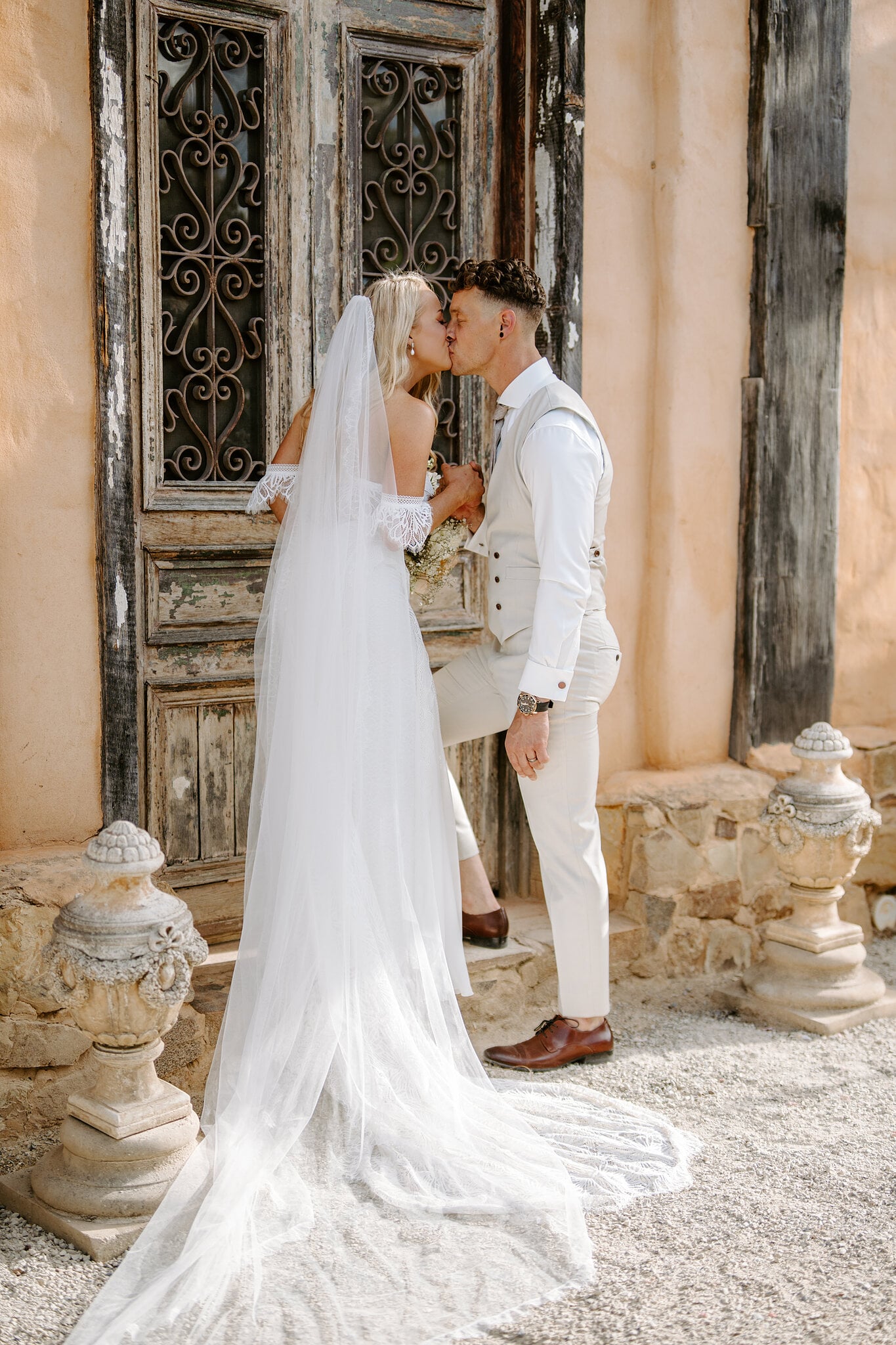 Samantha and Harrison kiss at the weathered carved doors between two stone urns.