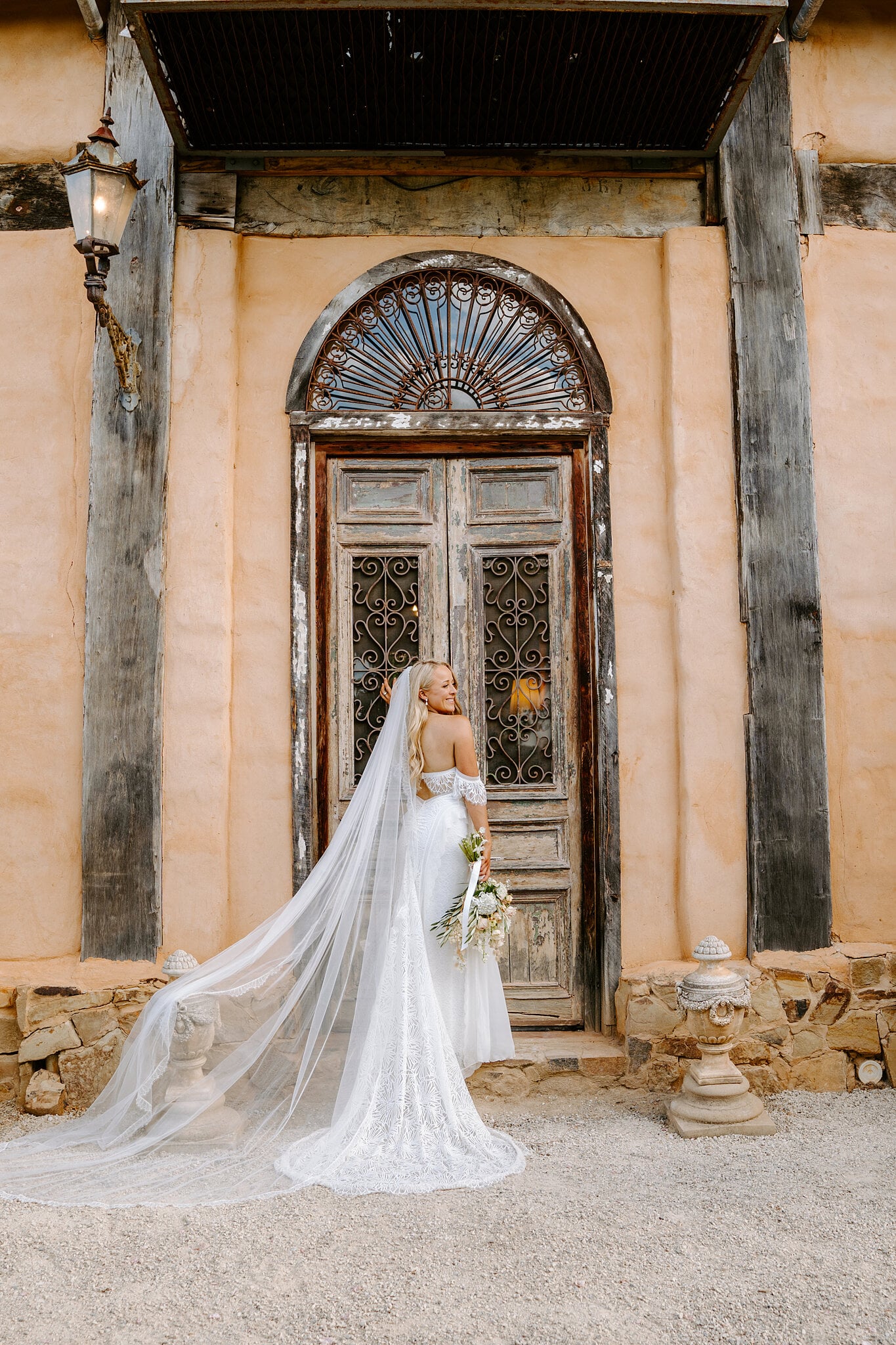 Samantha looks back from the arched door with its iron fanlight and lantern, her veil spread wide.