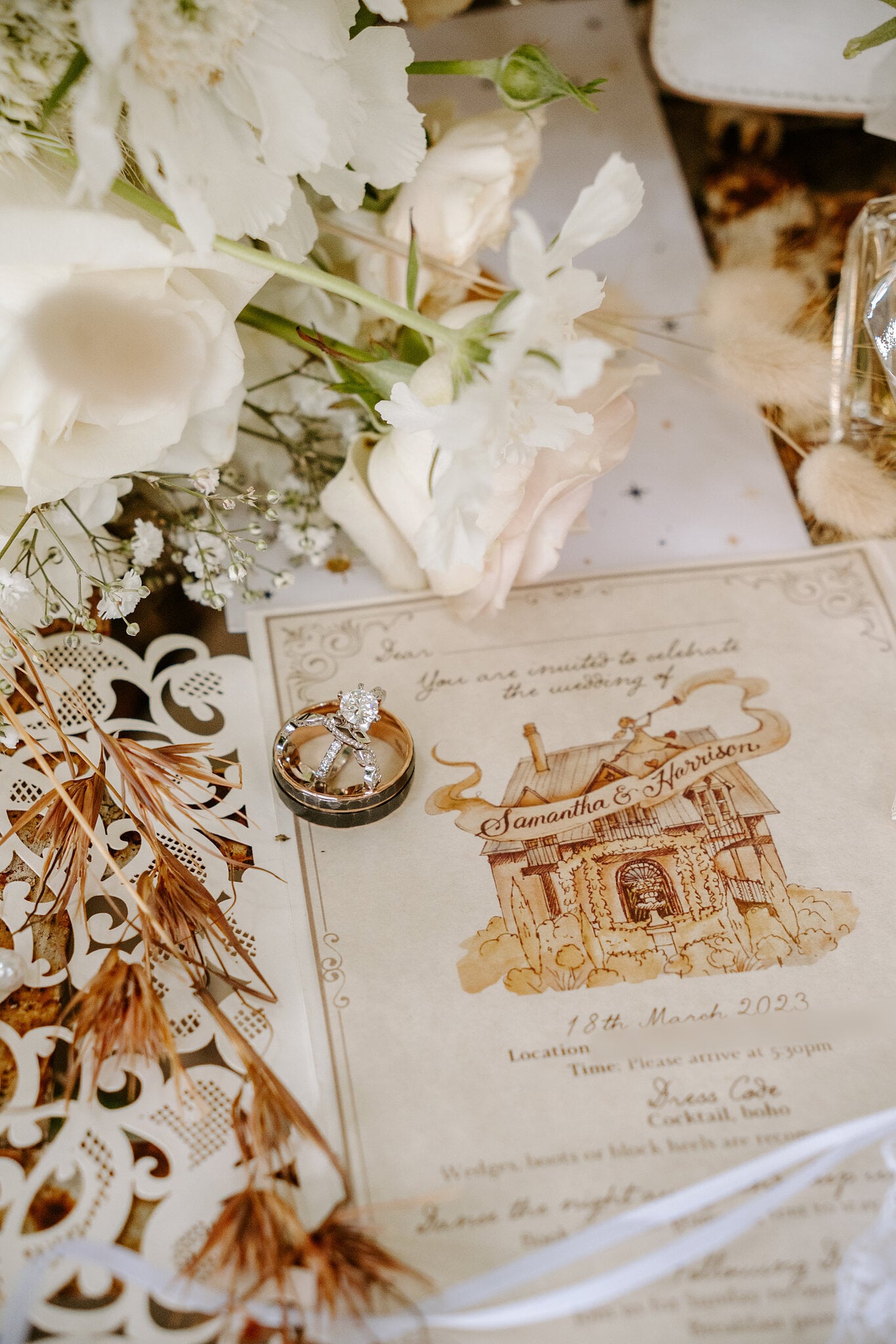 Wedding rings rest on an illustrated invitation among white flowers and dried grasses.