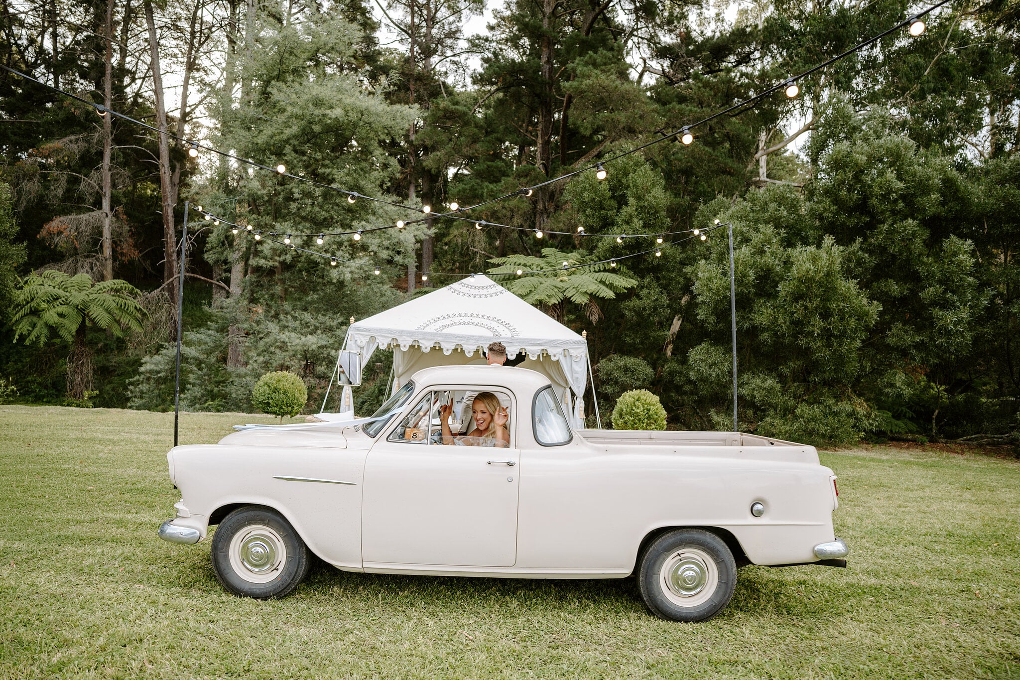 Samantha waves from the cream 1959 Holden ute under festoon lights at the reception.