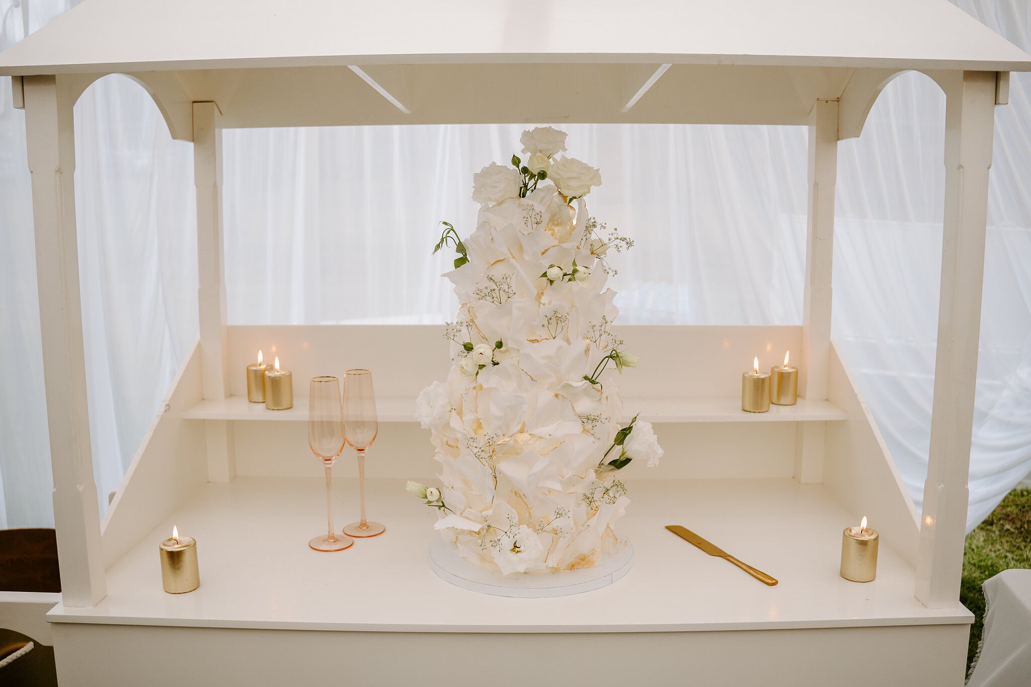 A tall white petal wedding cake in a candlelit timber hutch with two rose gold flutes.