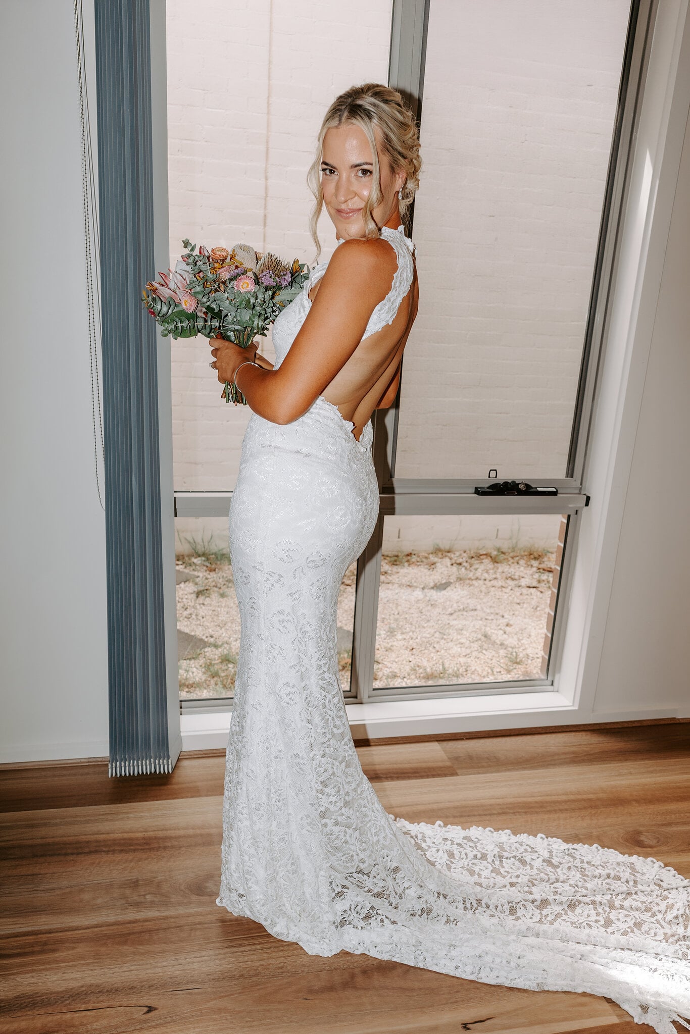 Emma glances back in her open-backed lace gown by the window, bouquet in hand.