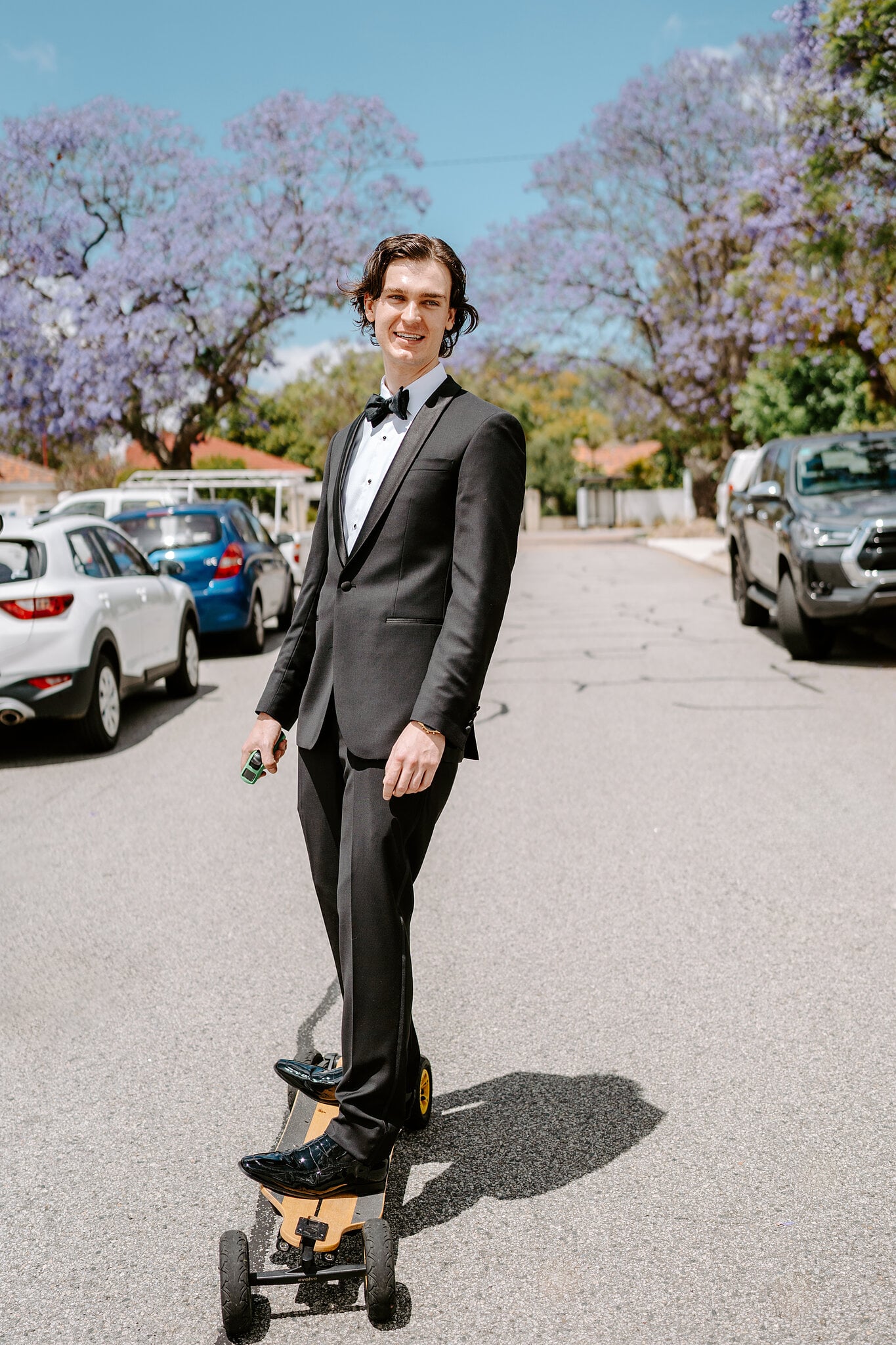George rides an electric skateboard down a jacaranda-lined street in his tuxedo, grinning.