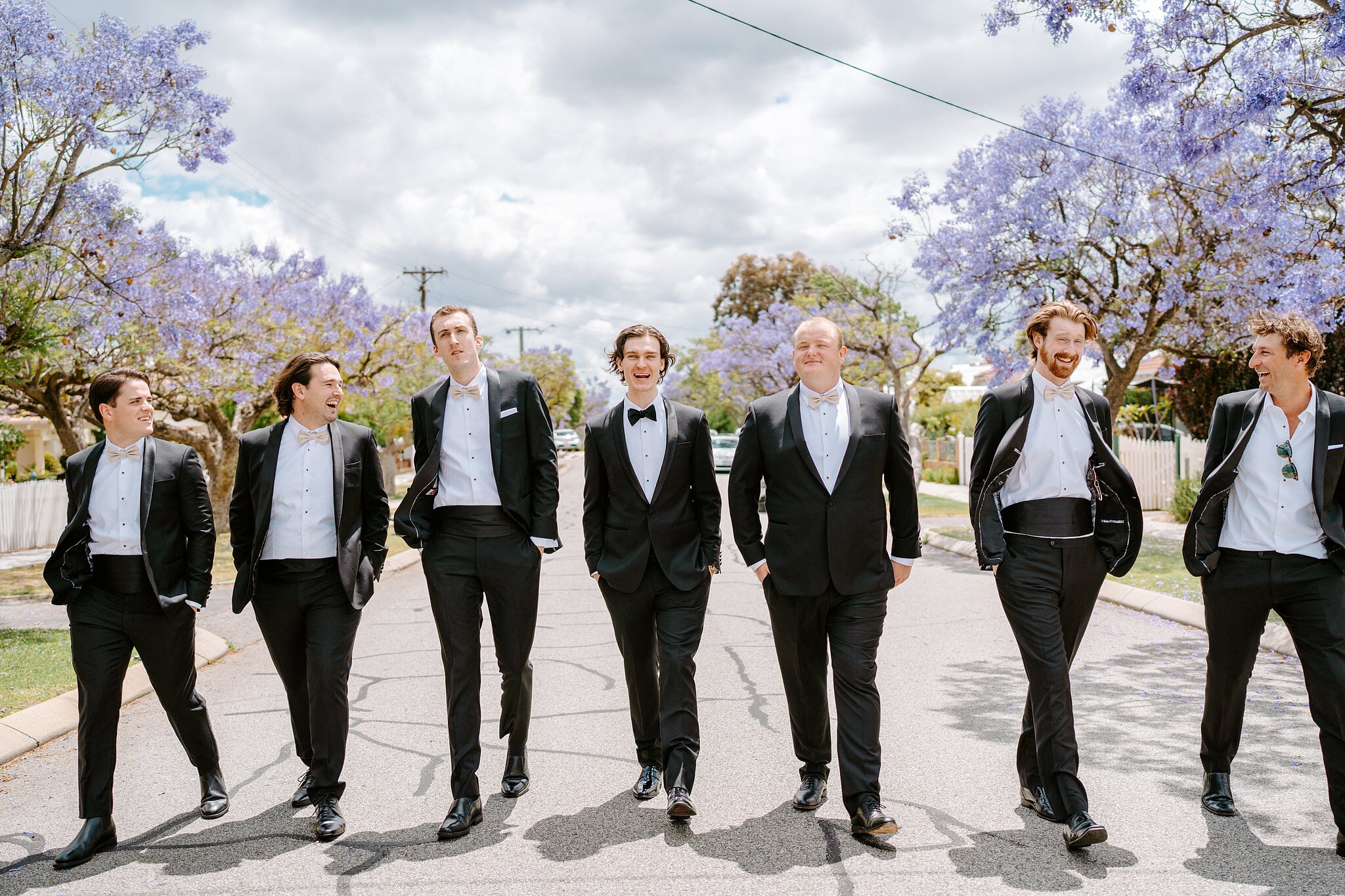 George and his six groomsmen stride down the street under purple jacaranda trees, laughing.