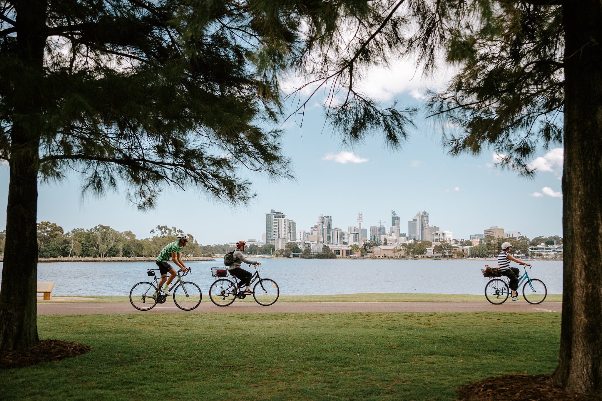 Cyclists roll along the riverside path under the pines, Perth city towers across the water.