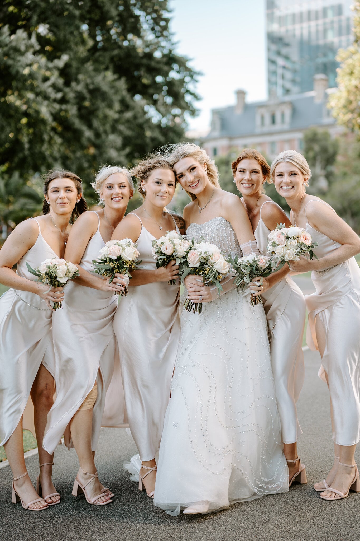 Claire and her five bridesmaids in champagne satin slip dresses, bouquets up, all smiles.
