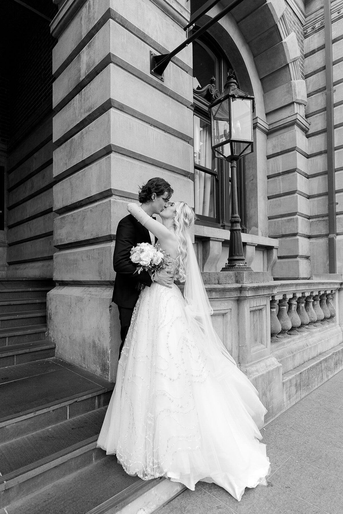 Black and white of Claire and George kissing on sandstone steps beside an old lantern.