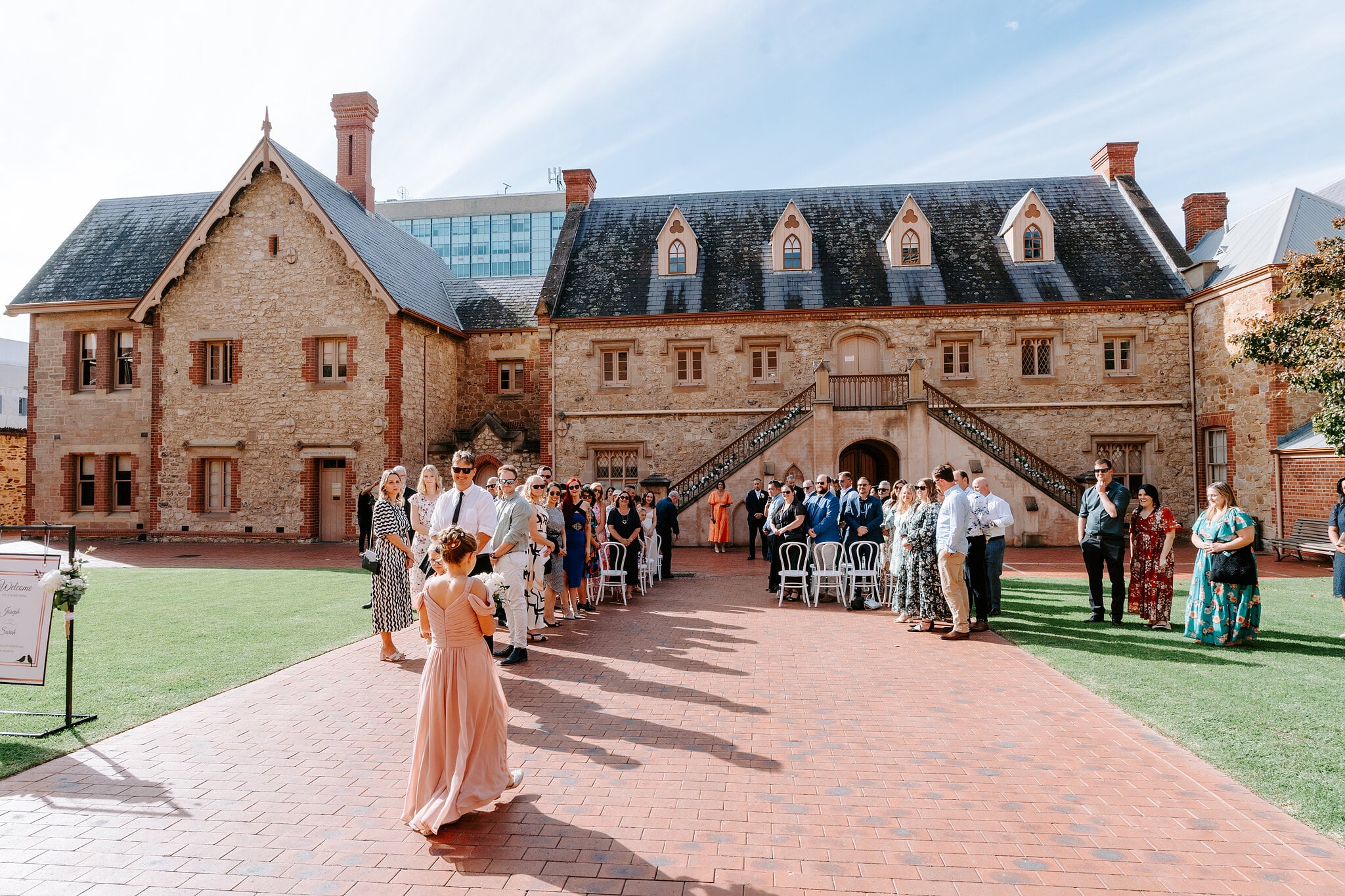 A bridesmaid in blush pink walks down the red-brick aisle as the guests stand.