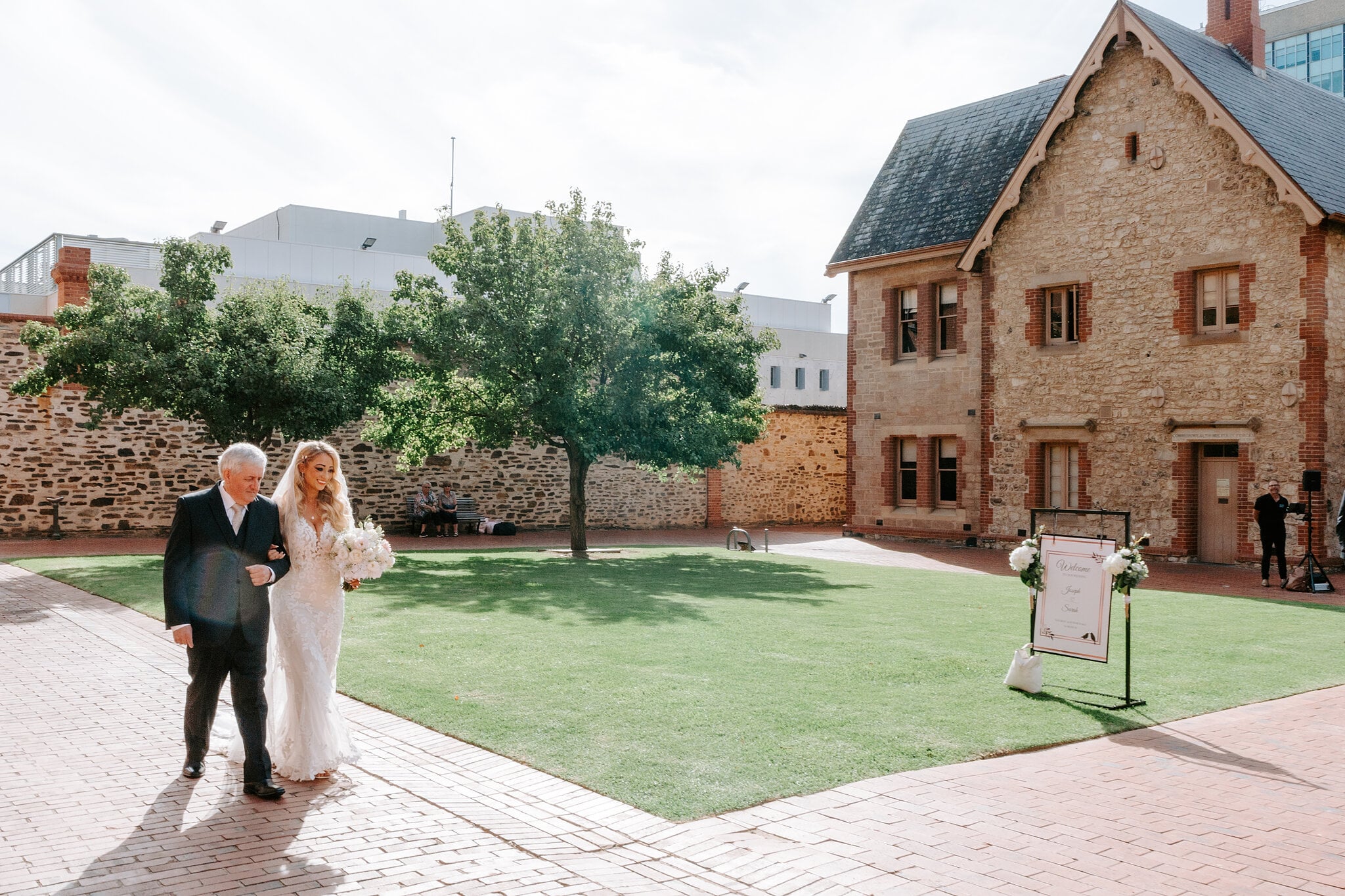 Sarah’s dad walks her arm in arm across the courtyard to her ceremony, bouquet in hand.