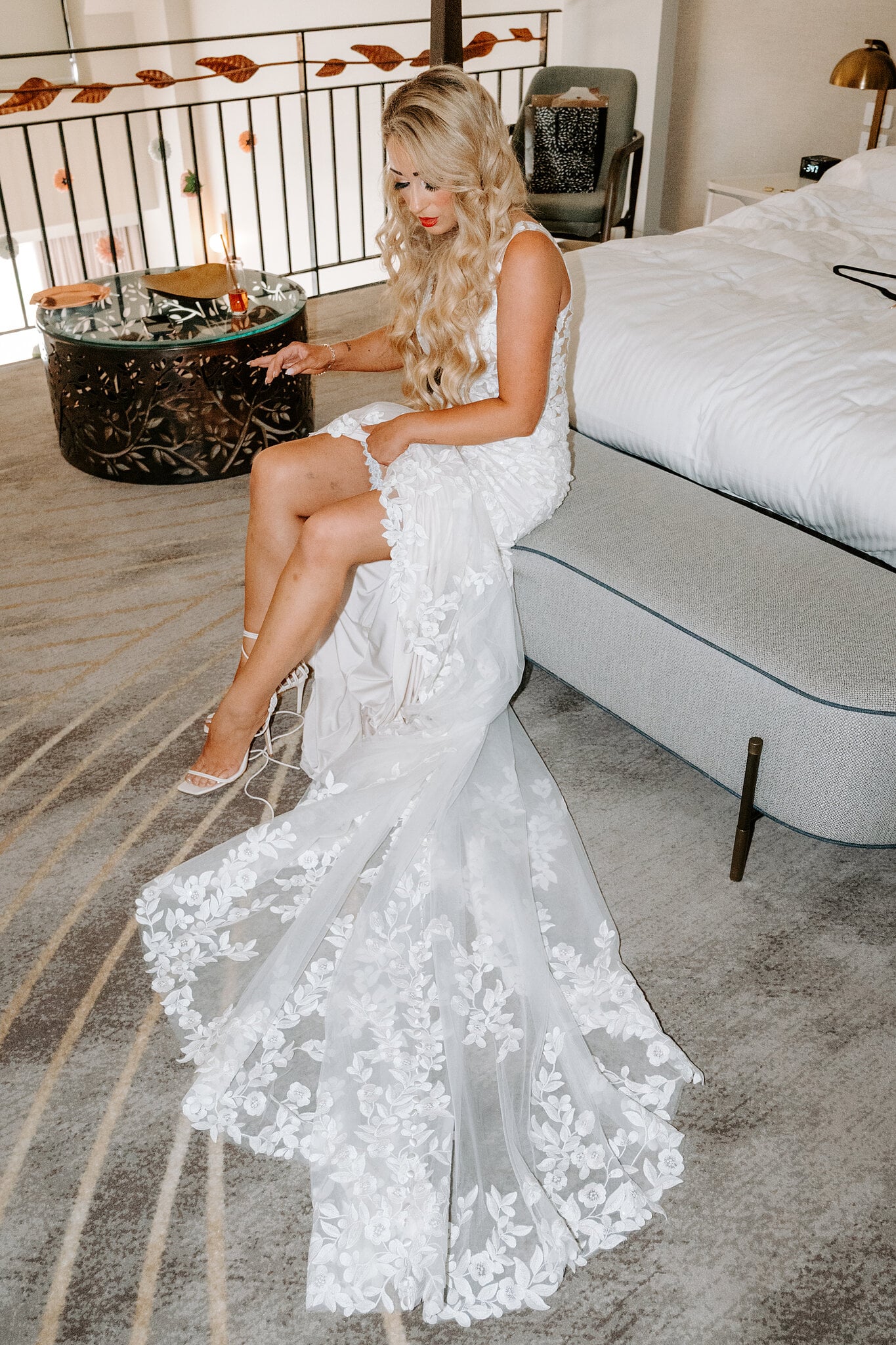 Sarah sits on the end of the bed in her lace mermaid gown, buckling her heels.