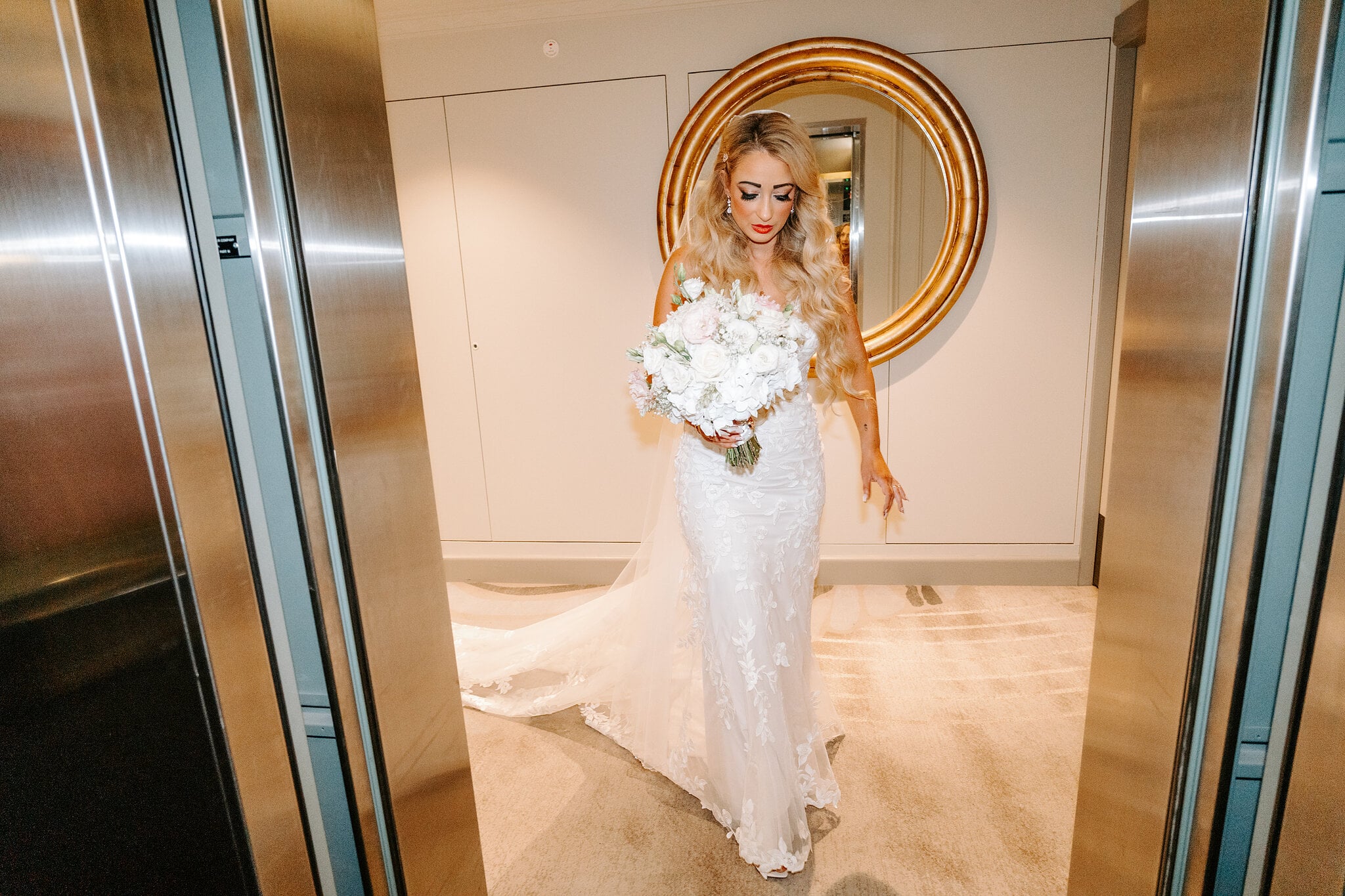 Sarah steps into the hotel lift with her white bouquet, a round gold mirror behind her.