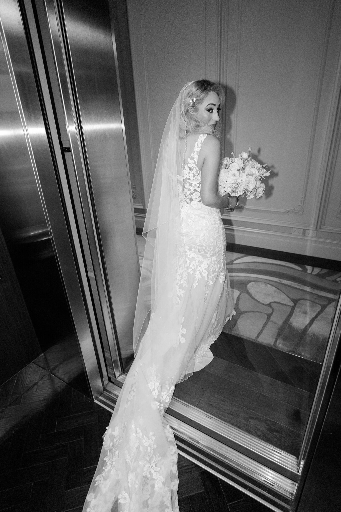 Sarah glances back over her shoulder in the lift, her veil and lace train behind her, in black and white.