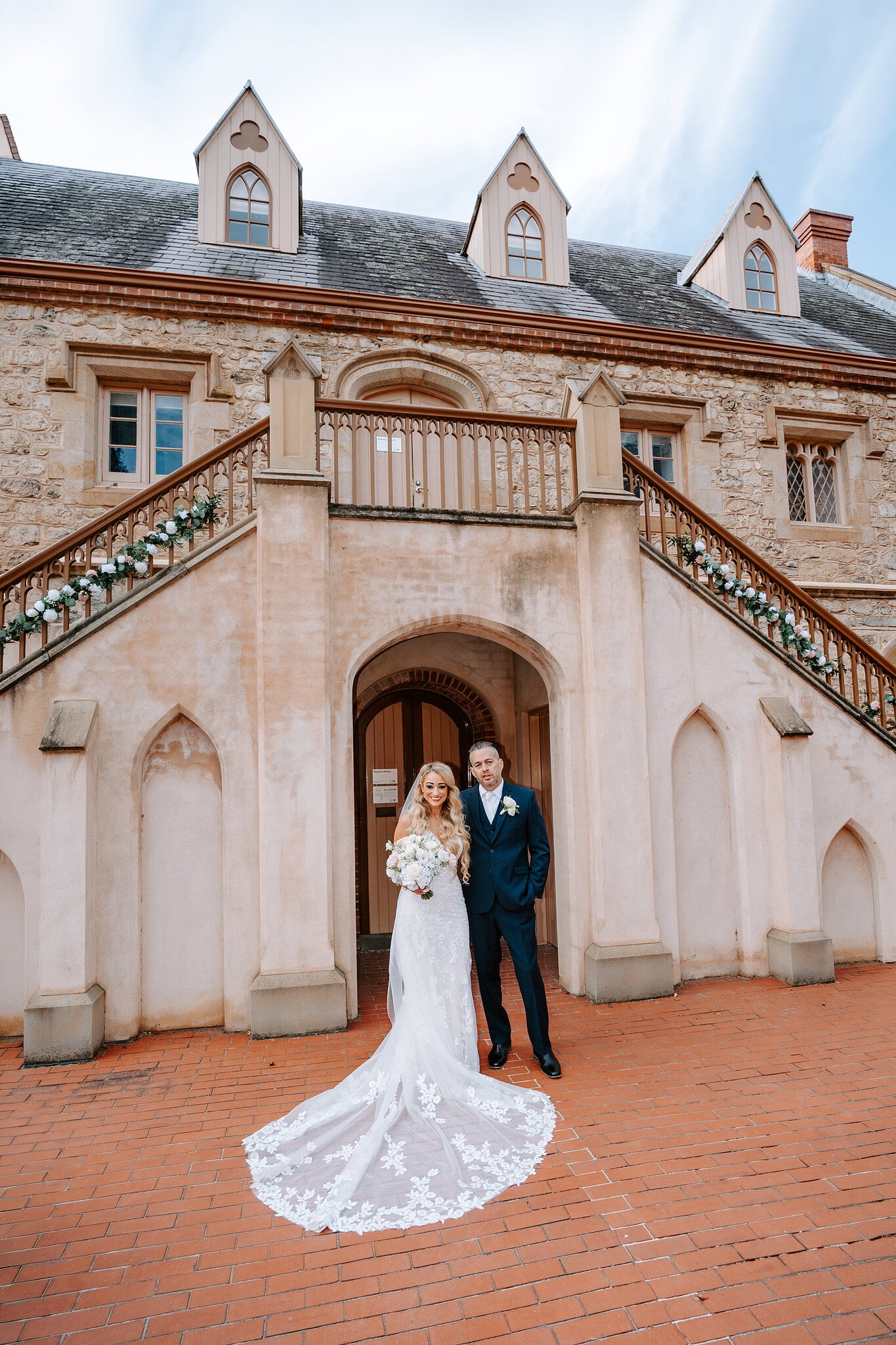 Sarah and Joe stand under the arch of the Armoury’s twin stone staircase, her lace train spread across the bricks.