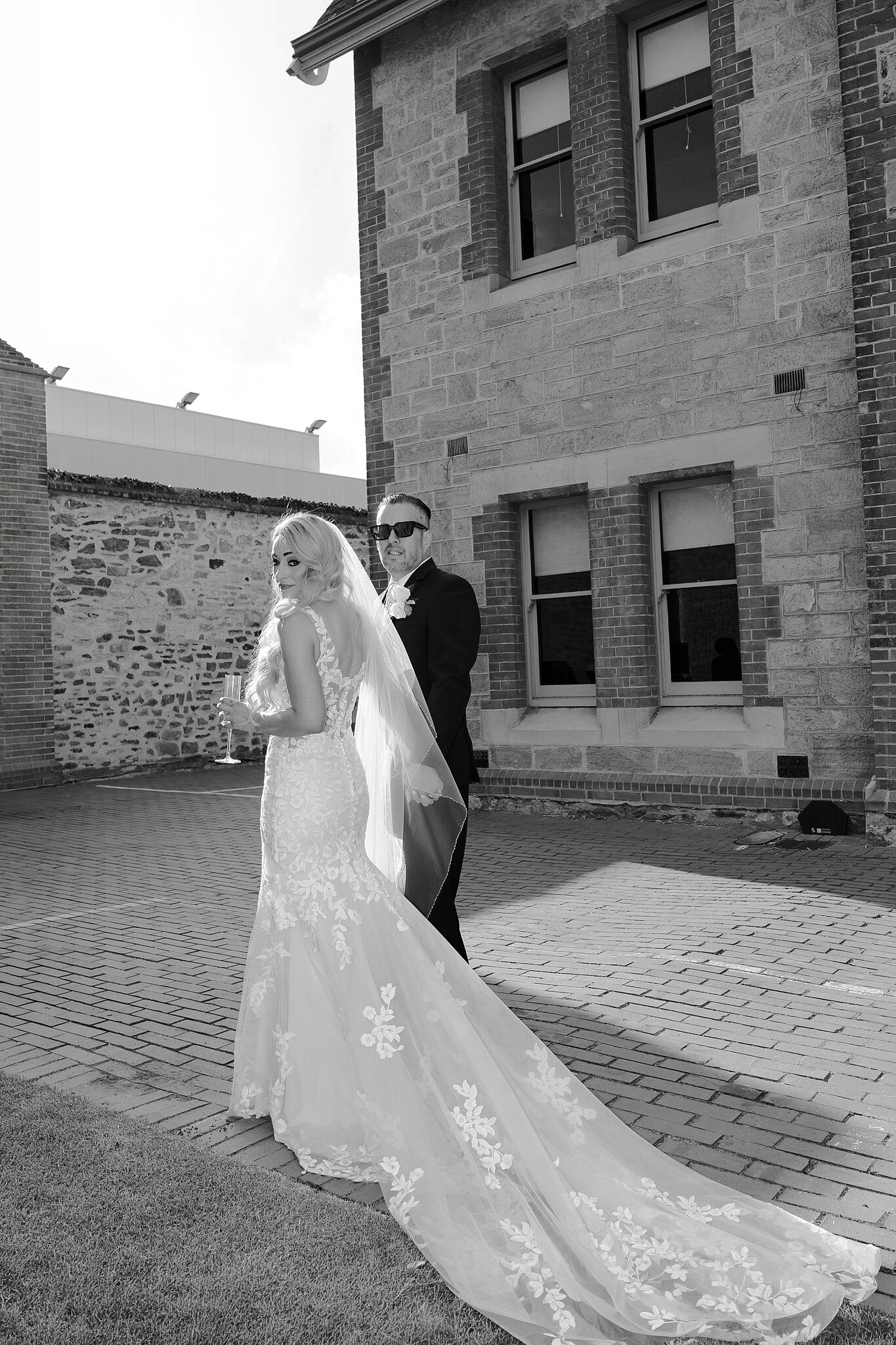 Sarah looks back with a glass of champagne as Joe waits in sunglasses beside the old stone buildings, in black and white.