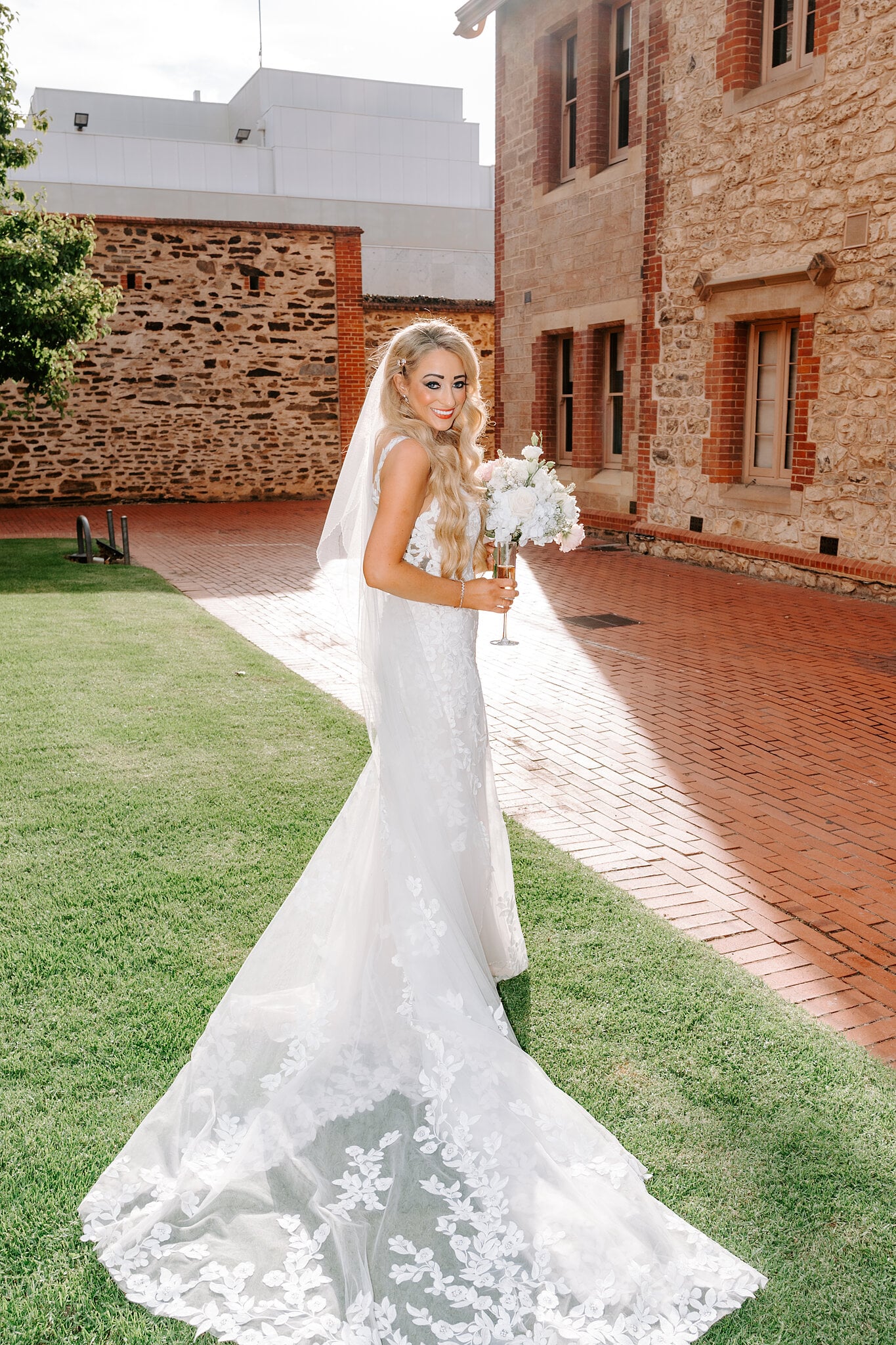 Sarah stands on the courtyard lawn with champagne and her bouquet, her lace train catching the sun.