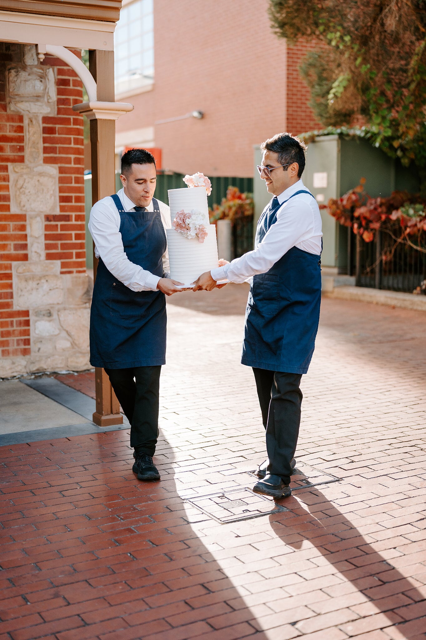 Two caterers carry the white wedding cake across the courtyard.