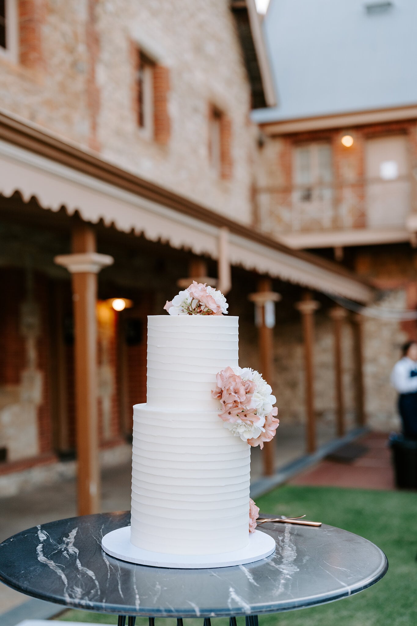 A white tiered wedding cake with blush flowers on a black marble table under the verandah.
