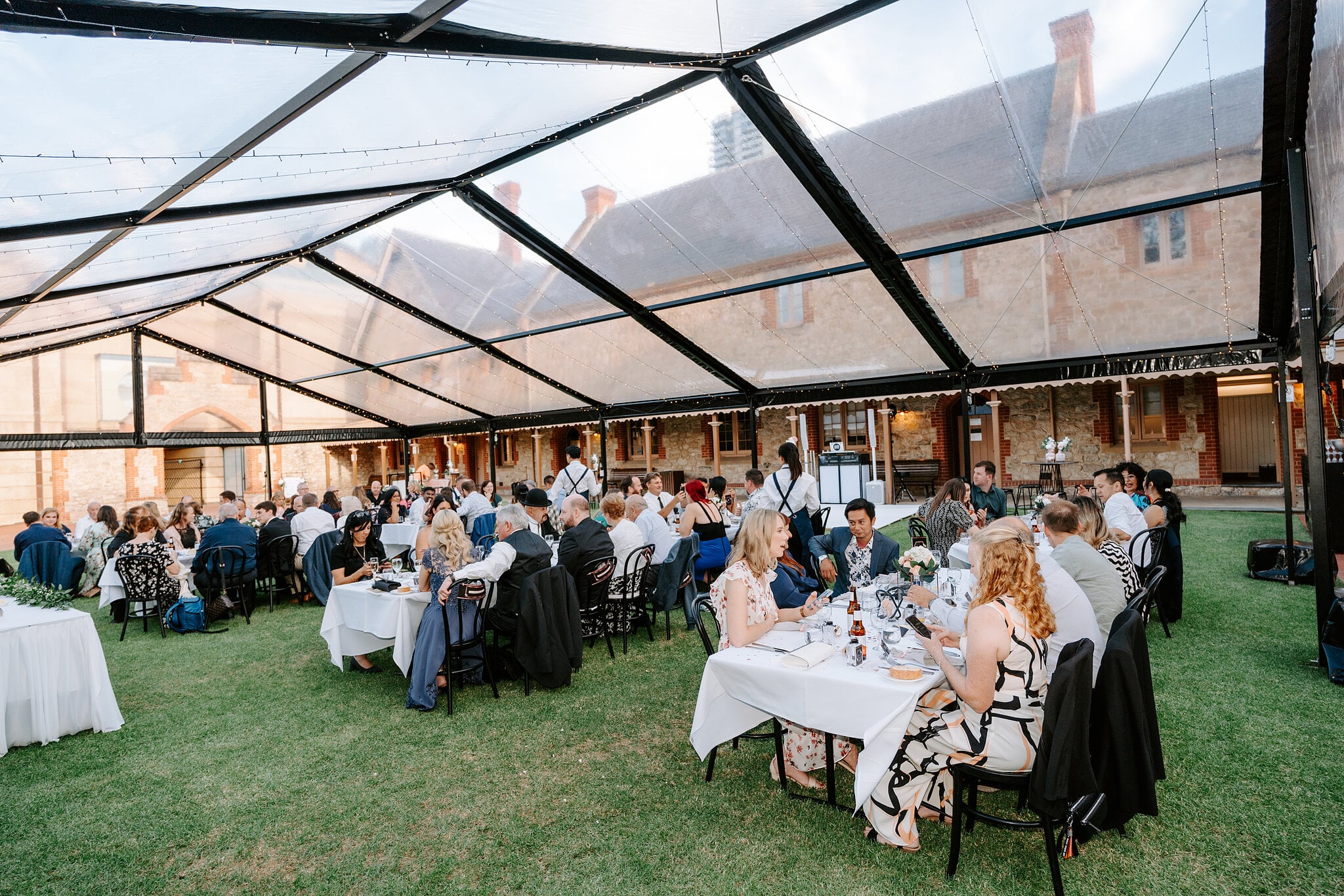 Guests dine at long tables under the clear marquee, the Armoury roofline overhead.