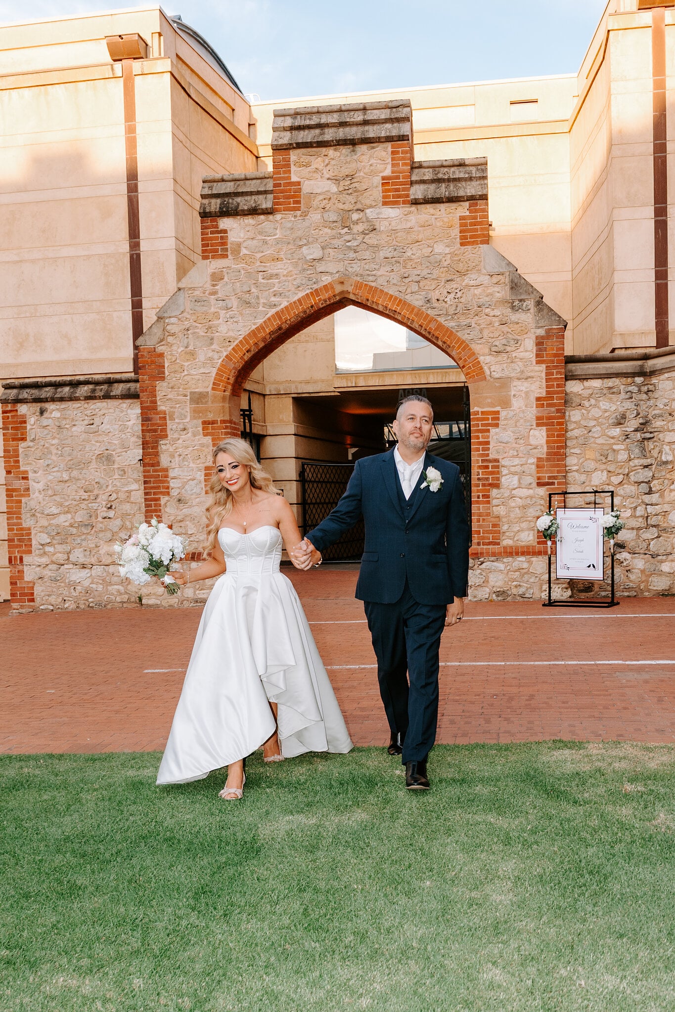 Sarah and Joe walk hand in hand from the stone archway onto the lawn, Sarah in a strapless satin gown.