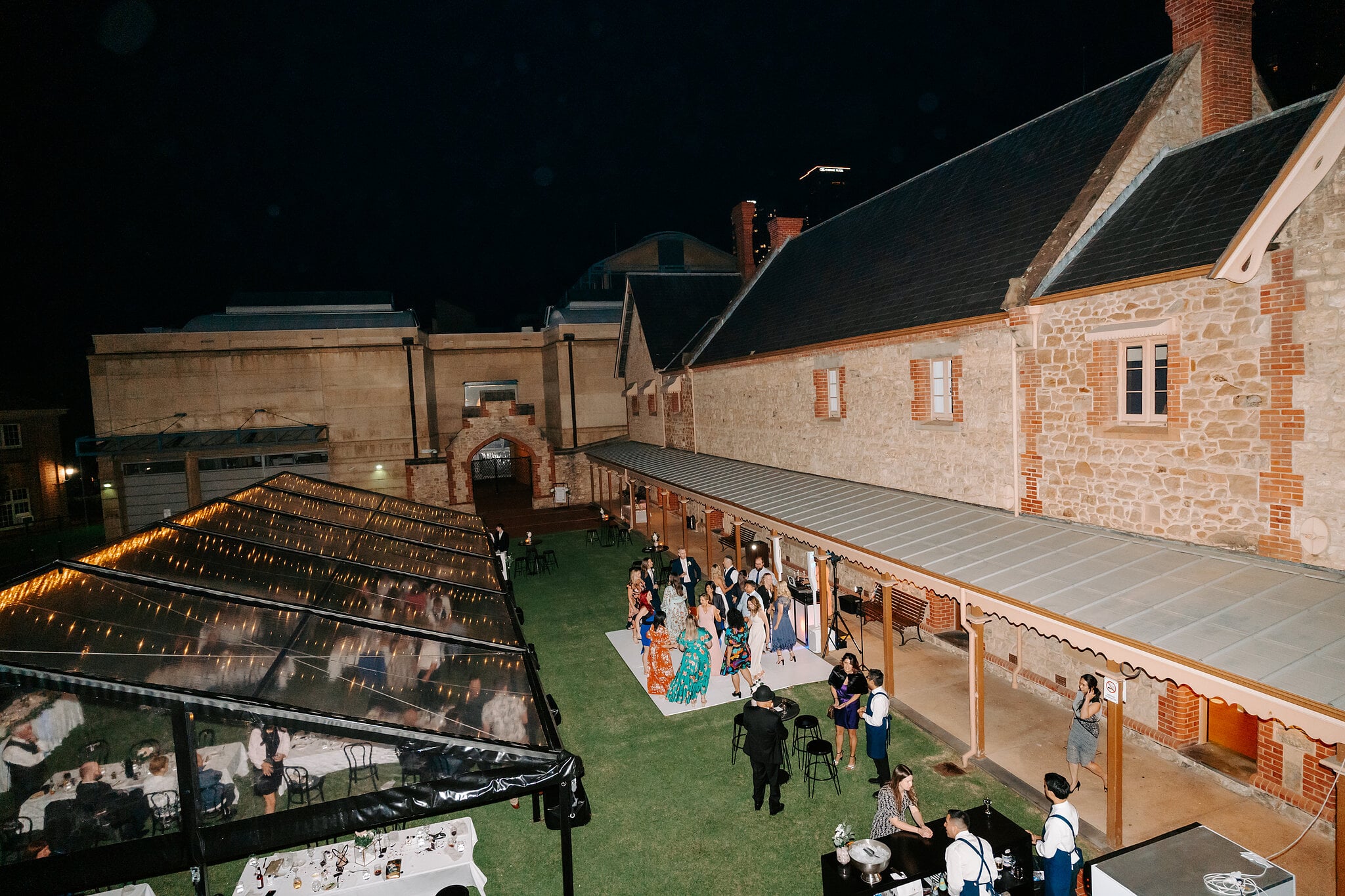 The clear marquee, lawn and dance floor beside the long stone building, seen from above at night.